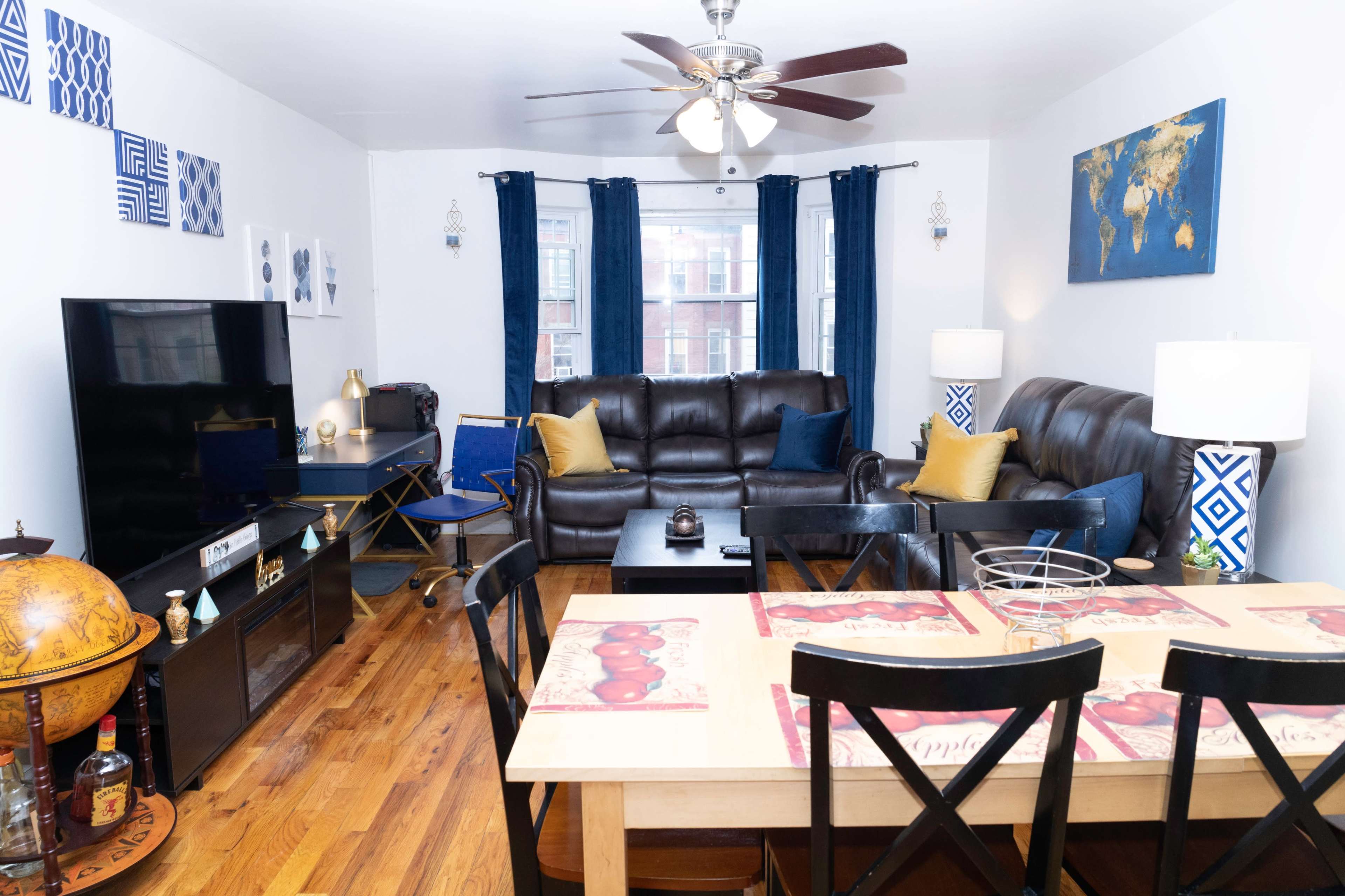 The living room features two brown leather sofas, a black television stand, a dining table with black chairs, and decor including blue curtains and a world map on the wall.