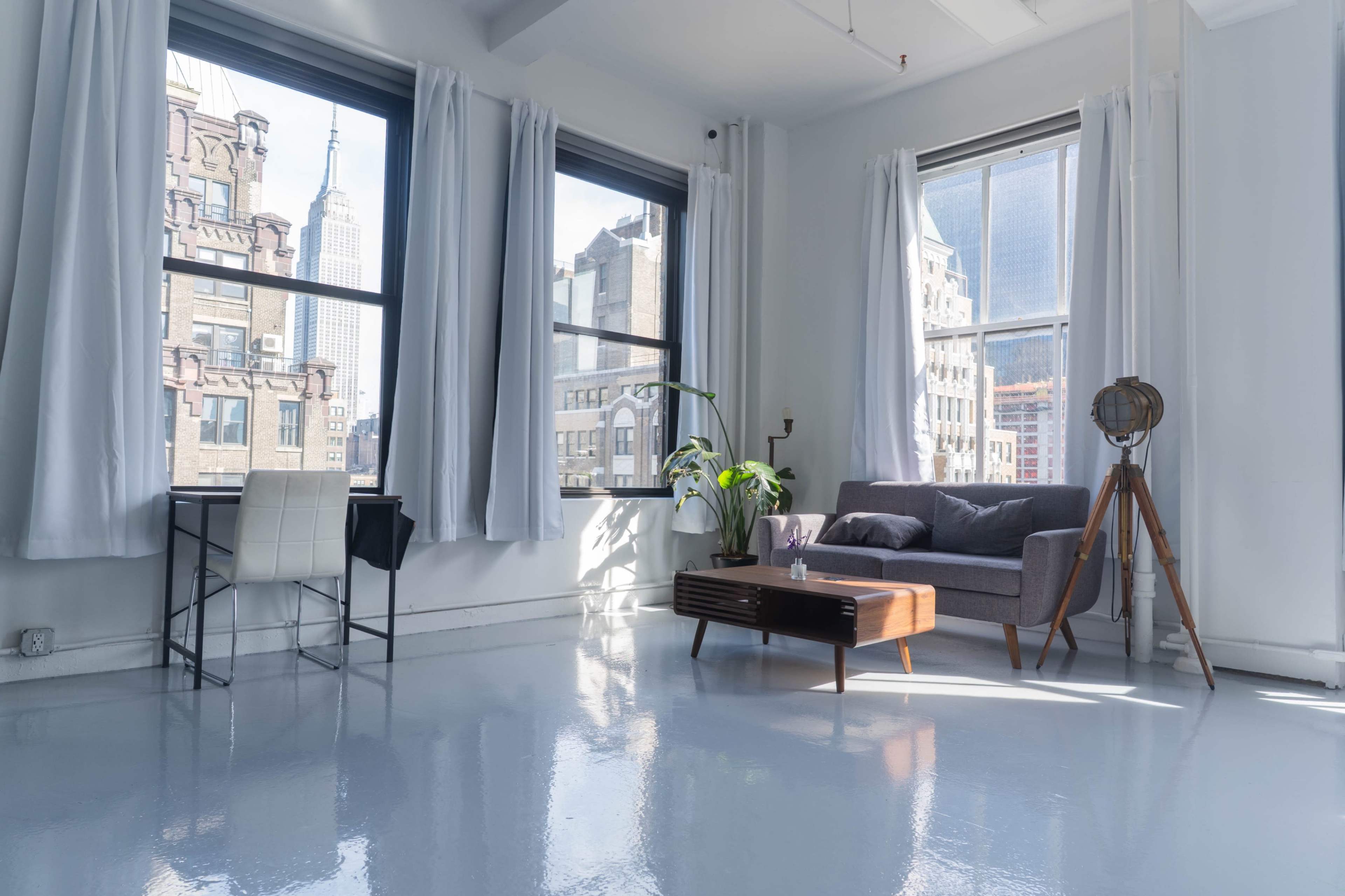 The image shows a bright, modern living space with large windows, minimalist furniture, and a view of city buildings.