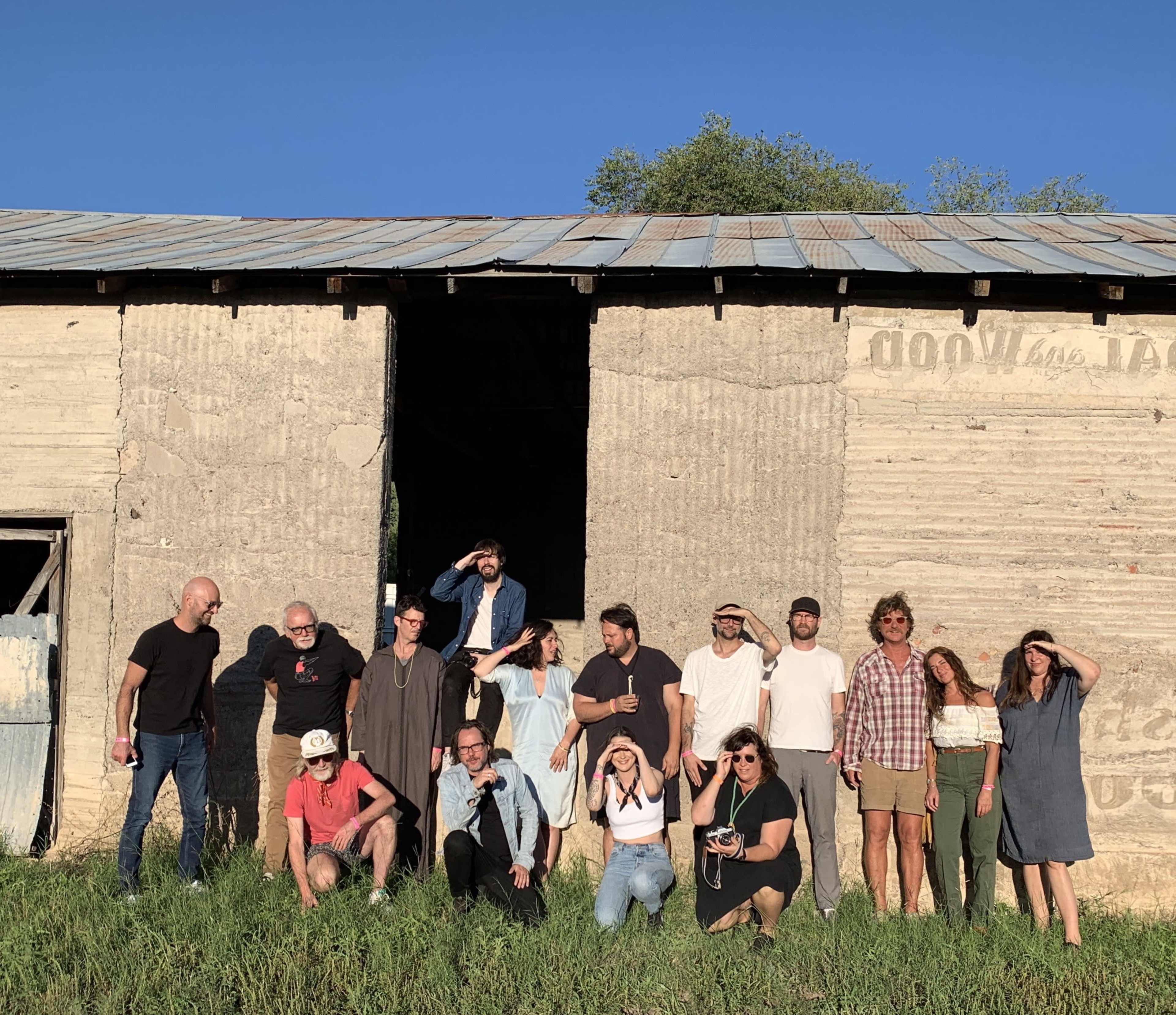 A group of people stands in front of a large, weathered building with a blue sky above.