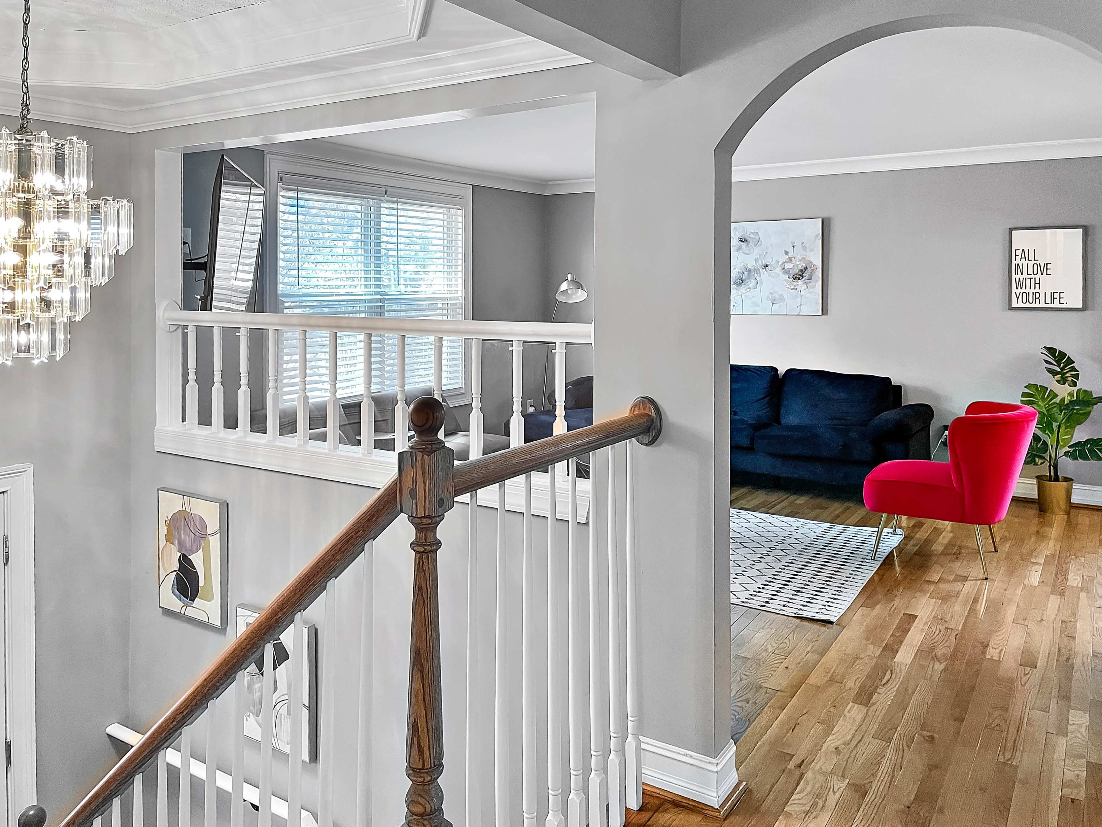 A staircase leading to a well-lit upper floor with a view of a living room featuring a dark sofa and a red accent chair.