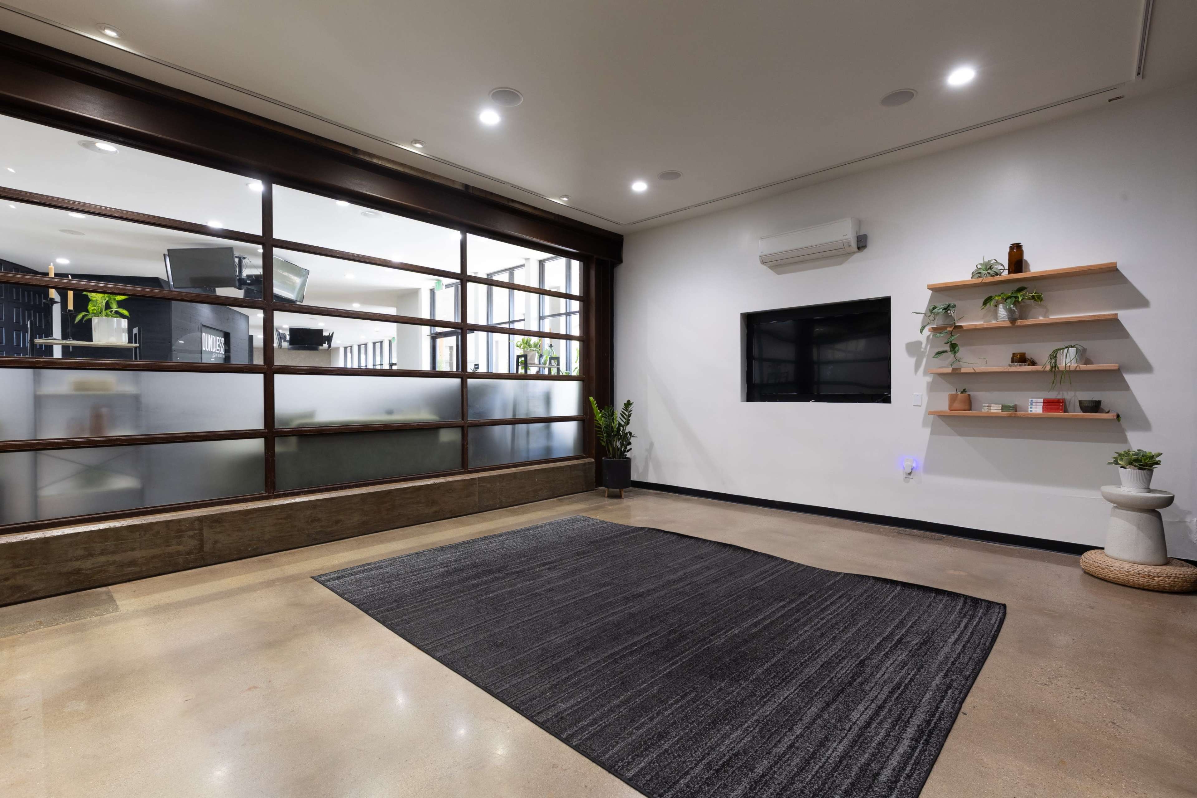 The image shows a modern office space featuring a wall-mounted television, a shelf with decorative items, and an area rug on a polished concrete floor.