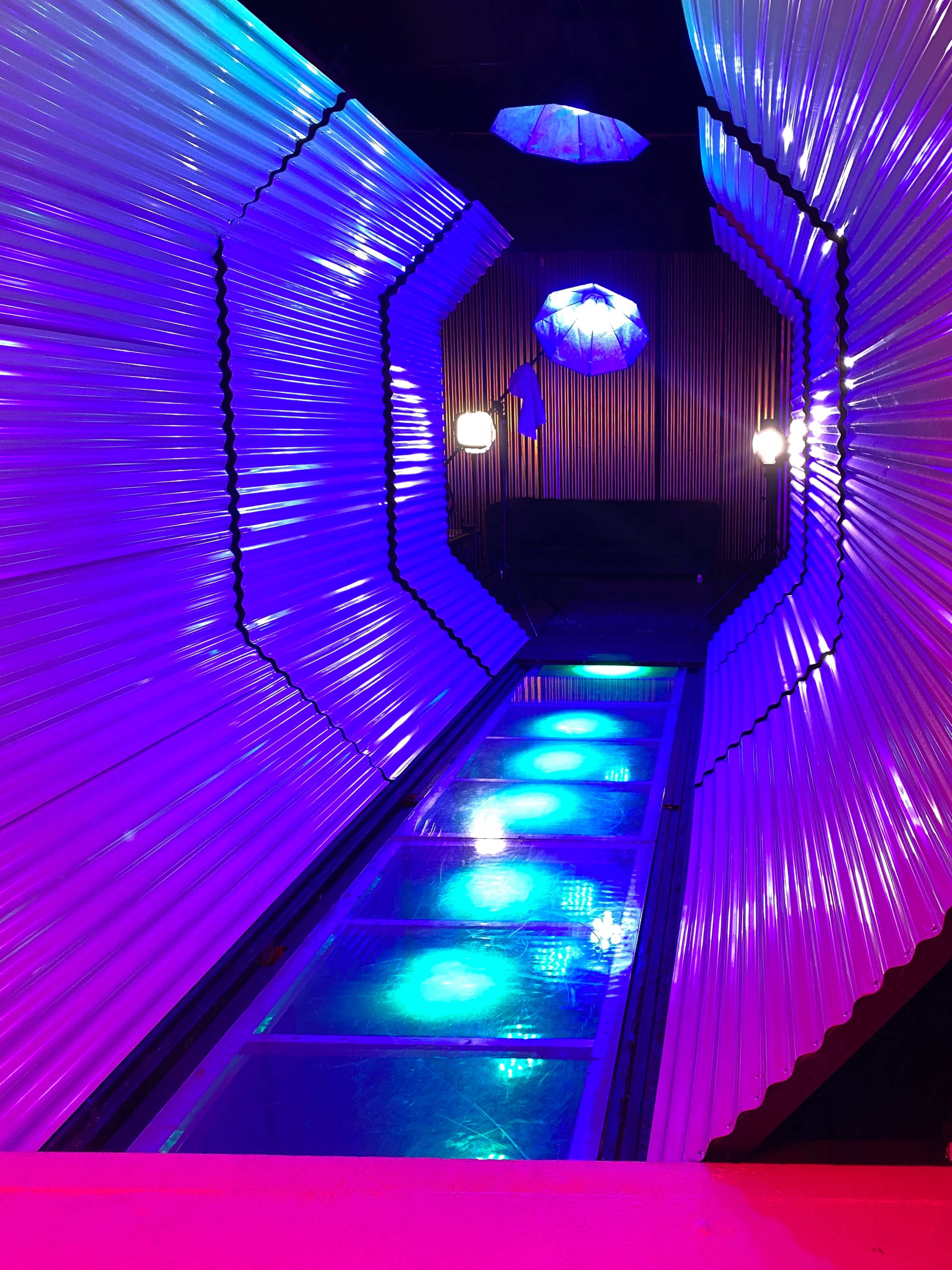A brightly lit, futuristic corridor lined with shiny metallic walls and illuminated by colorful lights.