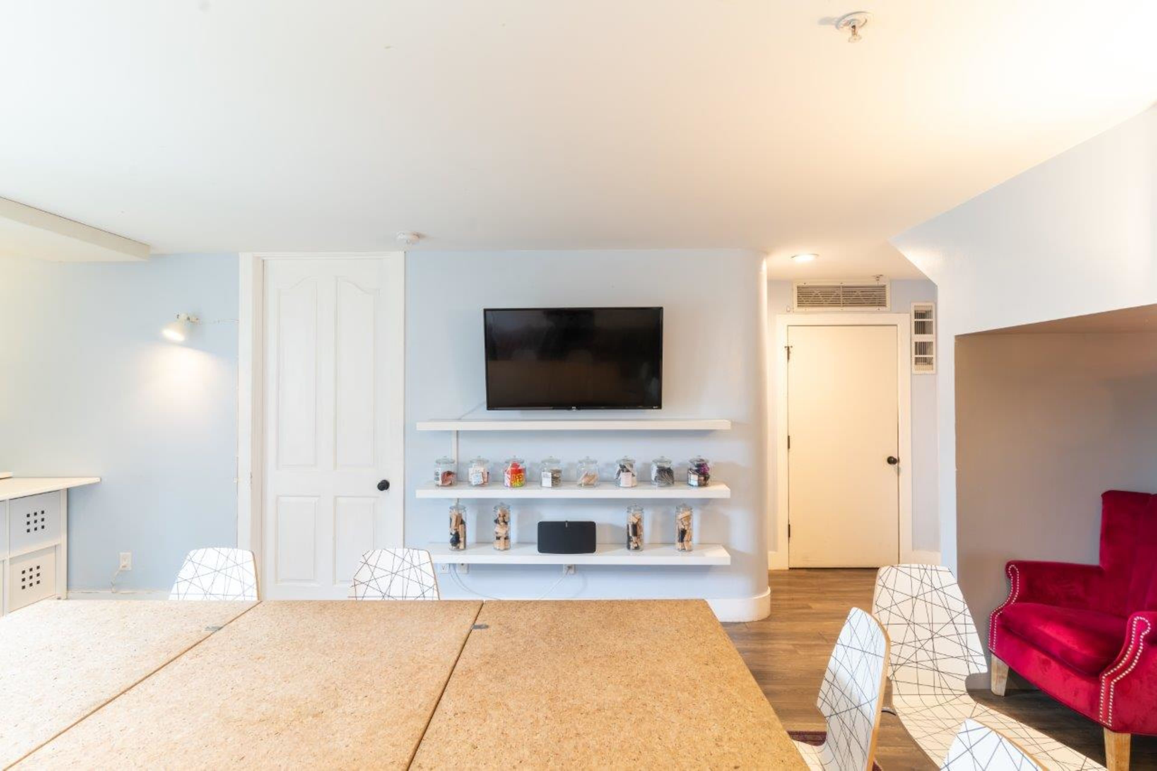 A modern meeting room with a large wooden table, a wall-mounted television, white shelves displaying jars, and a red armchair in the corner.