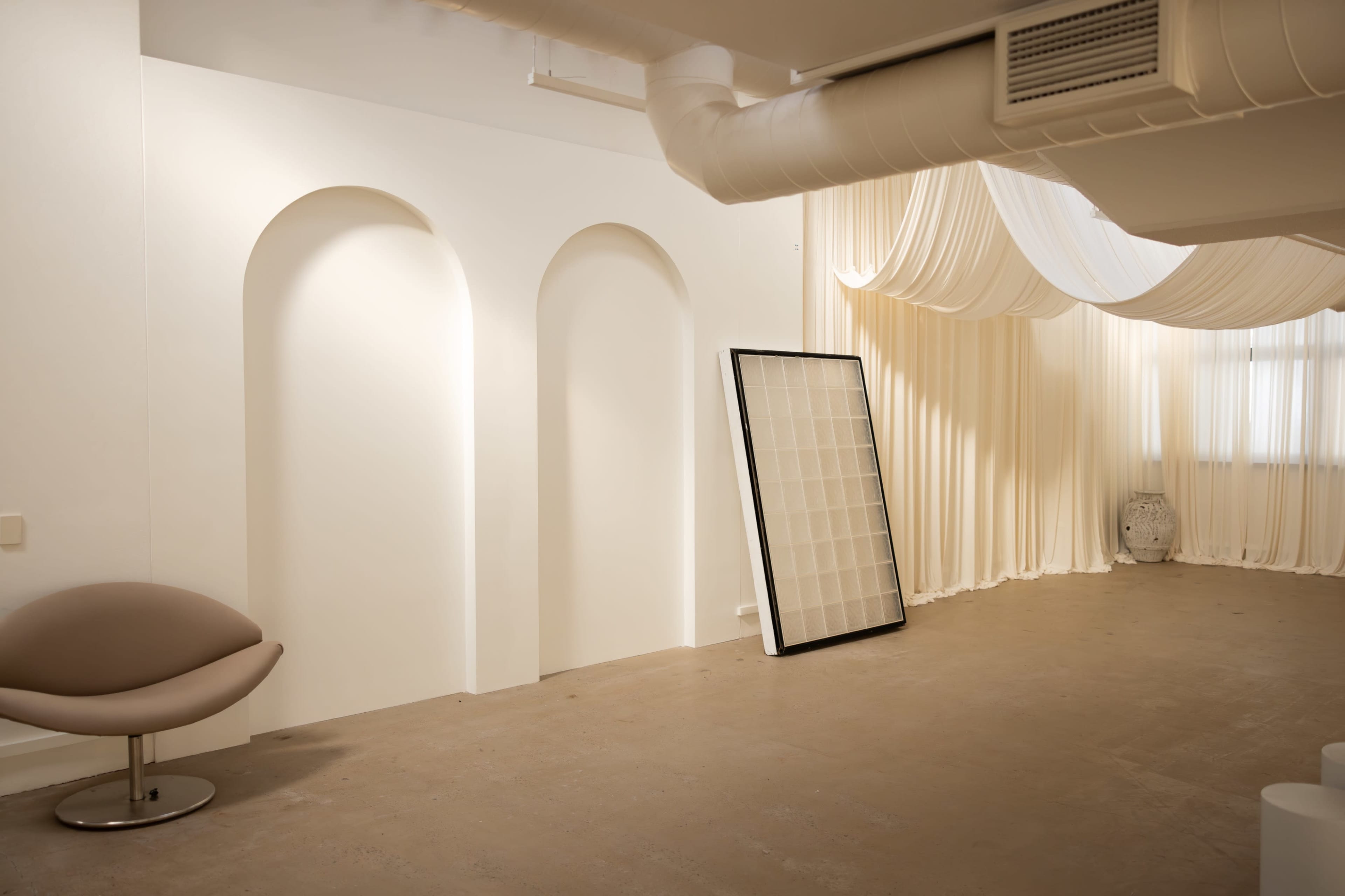 The image depicts a spacious interior room featuring curved wall arches, a large framed grid standing against the wall, and soft fabric draping from the ceiling.