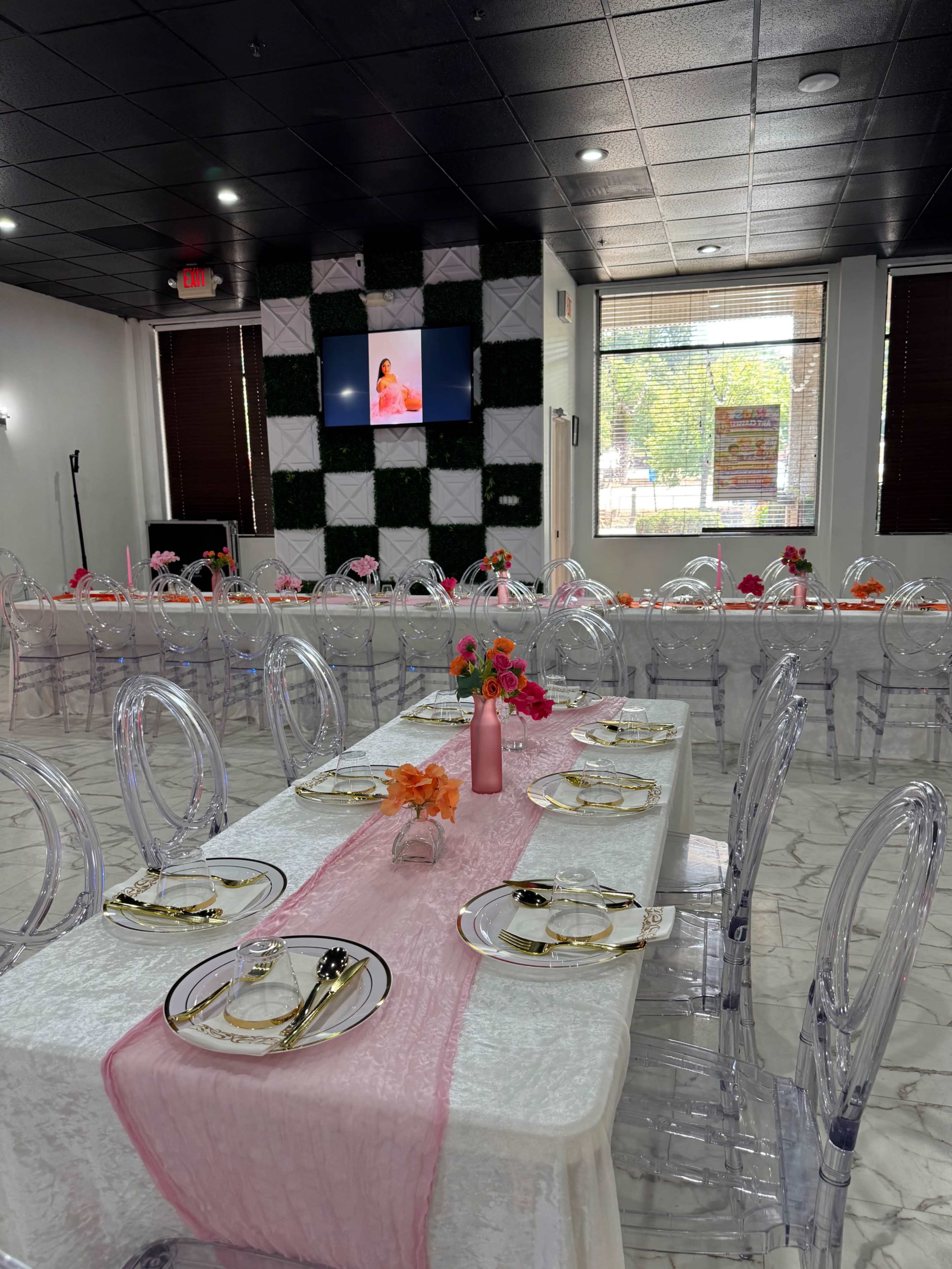 The image shows a dining area set up for an event, featuring long tables adorned with white tablecloths, clear chairs, and floral centerpieces, along with a wall-mounted screen in the background.