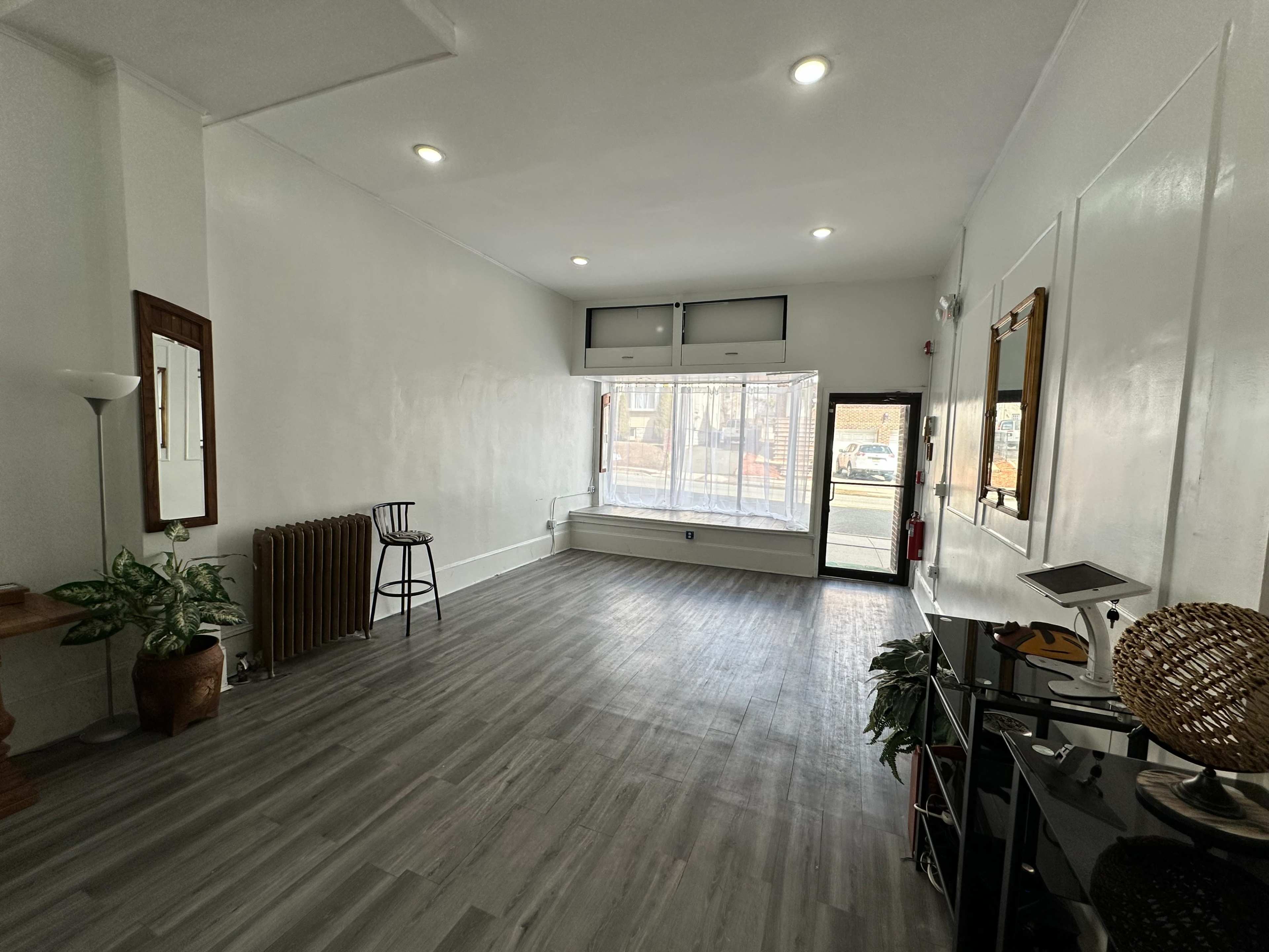 The image shows a sparsely furnished room with wooden flooring, a large window, and a few decorative items, including a plant and a mirror.