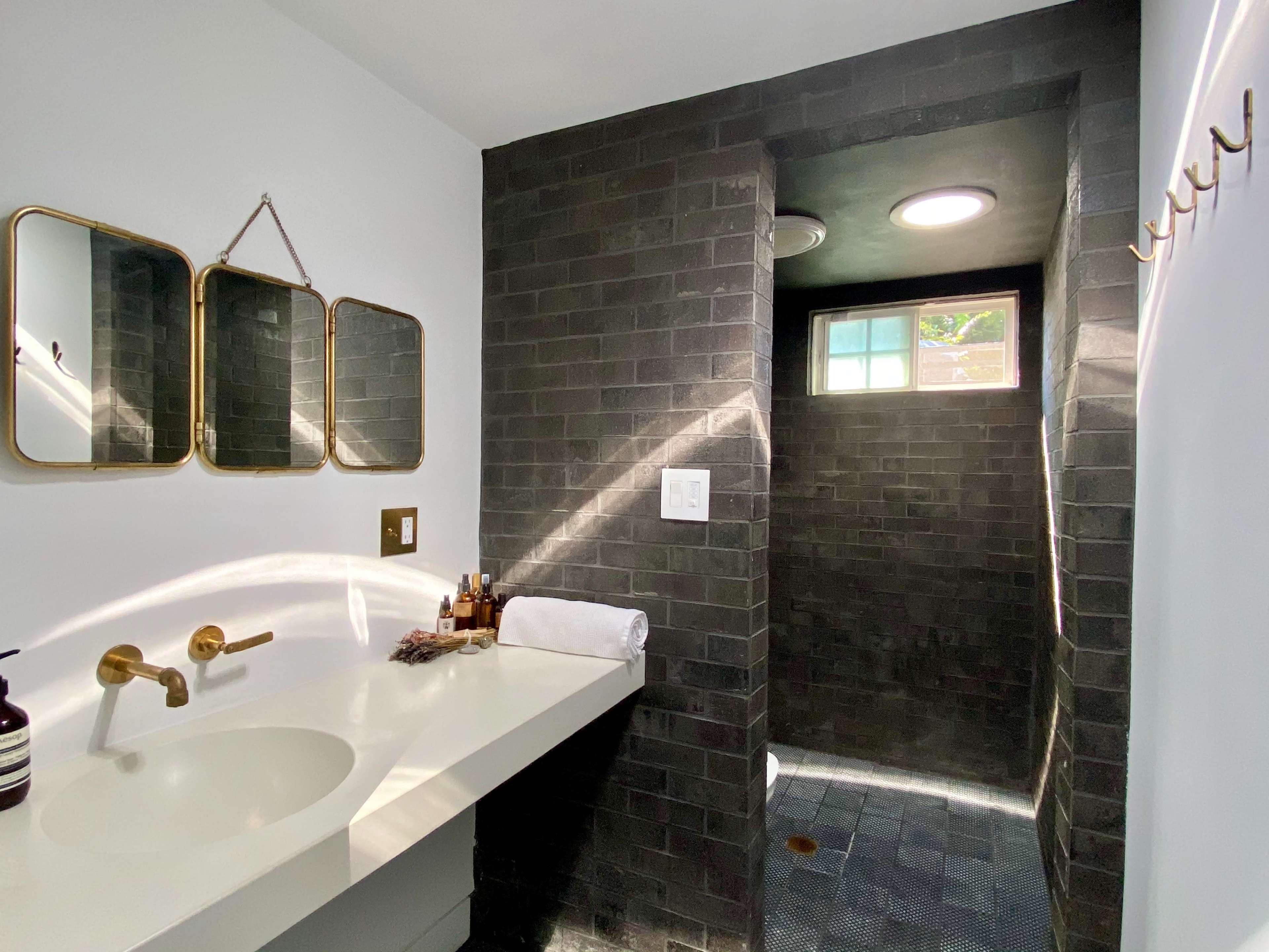 A modern bathroom with a double sink, three mirrors, a shower area with dark tiled walls, and natural light coming from a window.