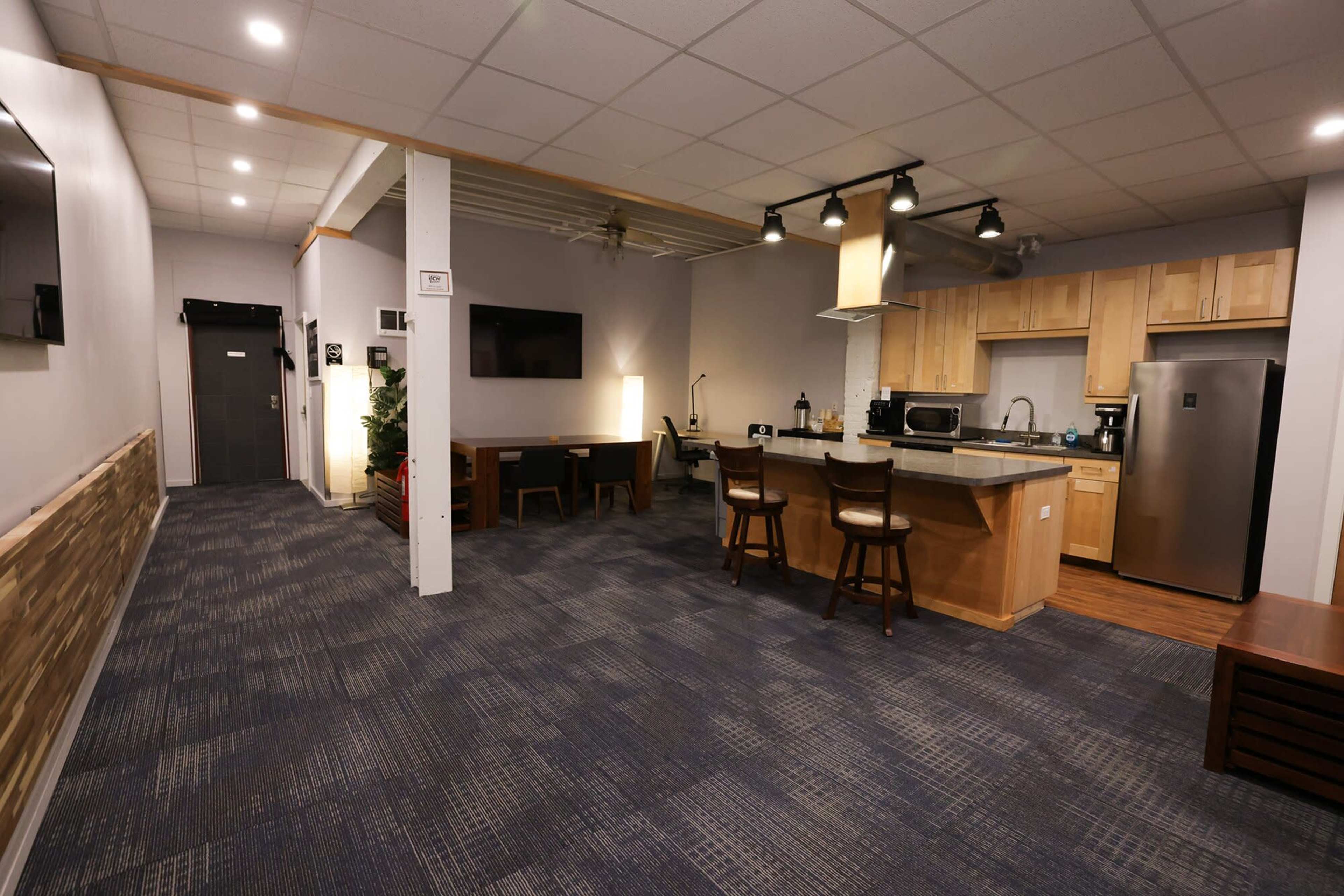 A modern kitchen and dining area with wood cabinetry, a countertop, and a small table with chairs, adjacent to a hallway.
