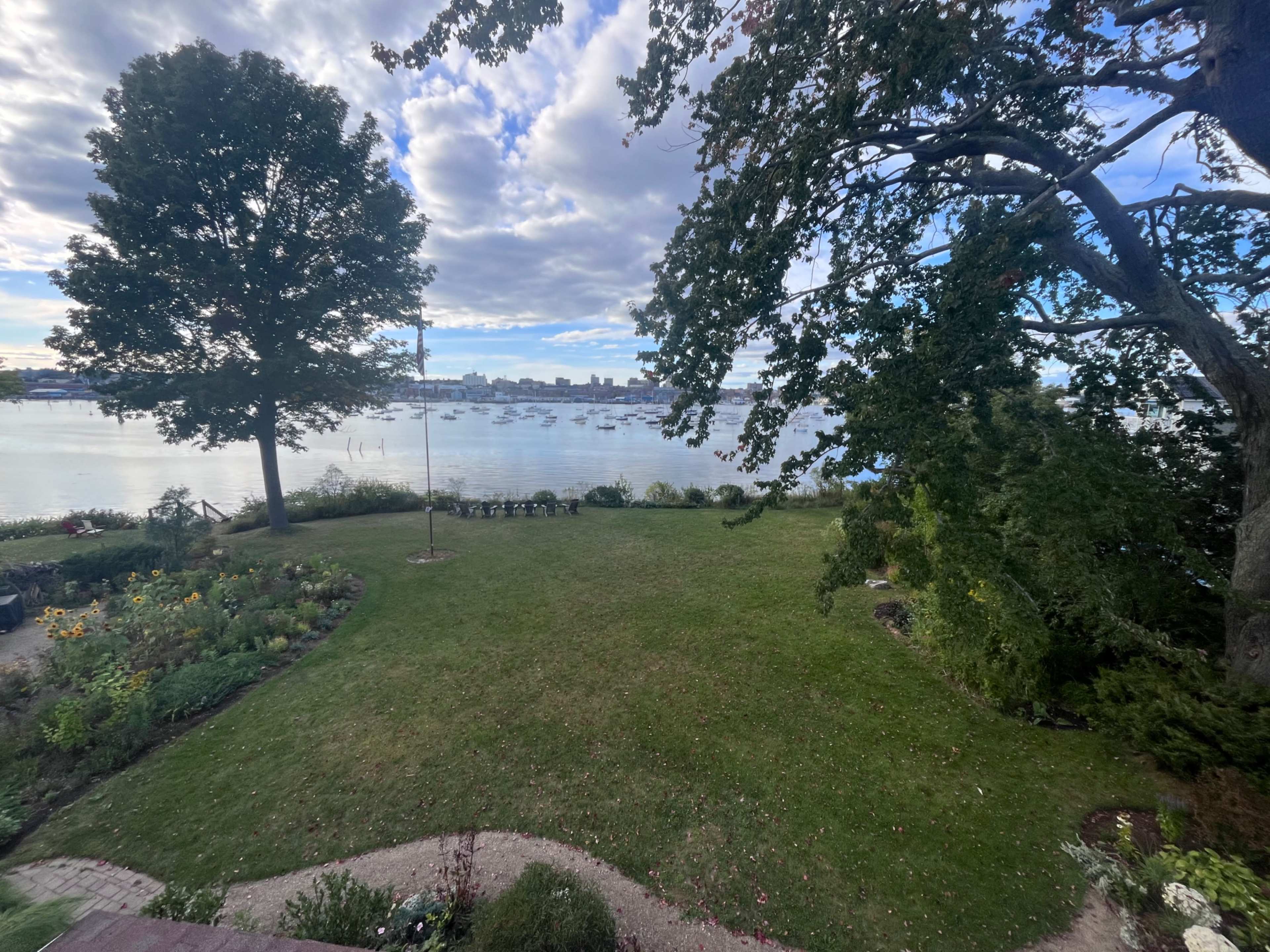 The image shows a grassy yard with flower beds, leading to a calm body of water dotted with boats and framed by trees under a cloudy sky.