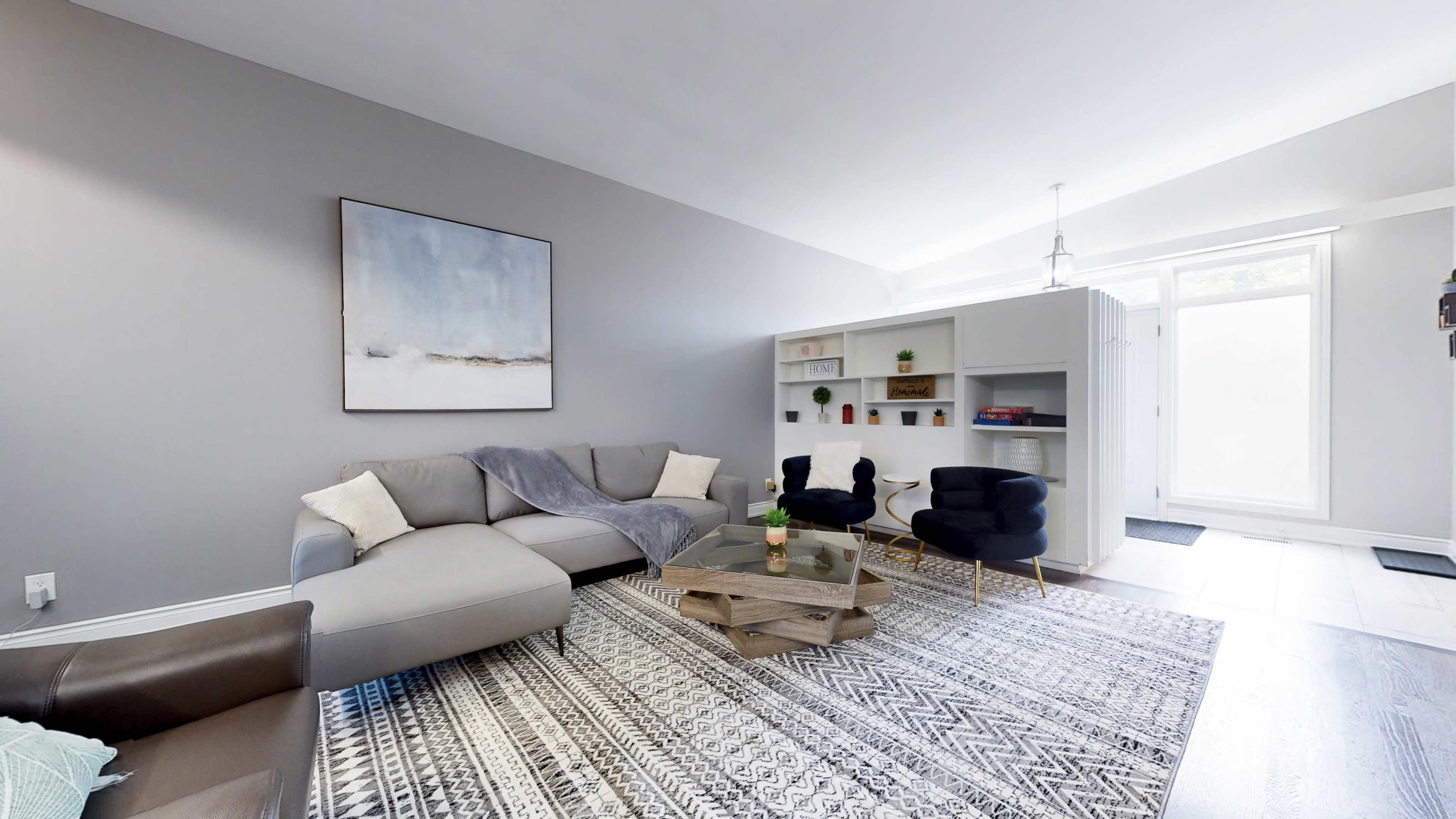 A modern living room features a gray sectional sofa, a wooden coffee table, and a bookshelf against a light wall, with large windows allowing natural light to enter.
