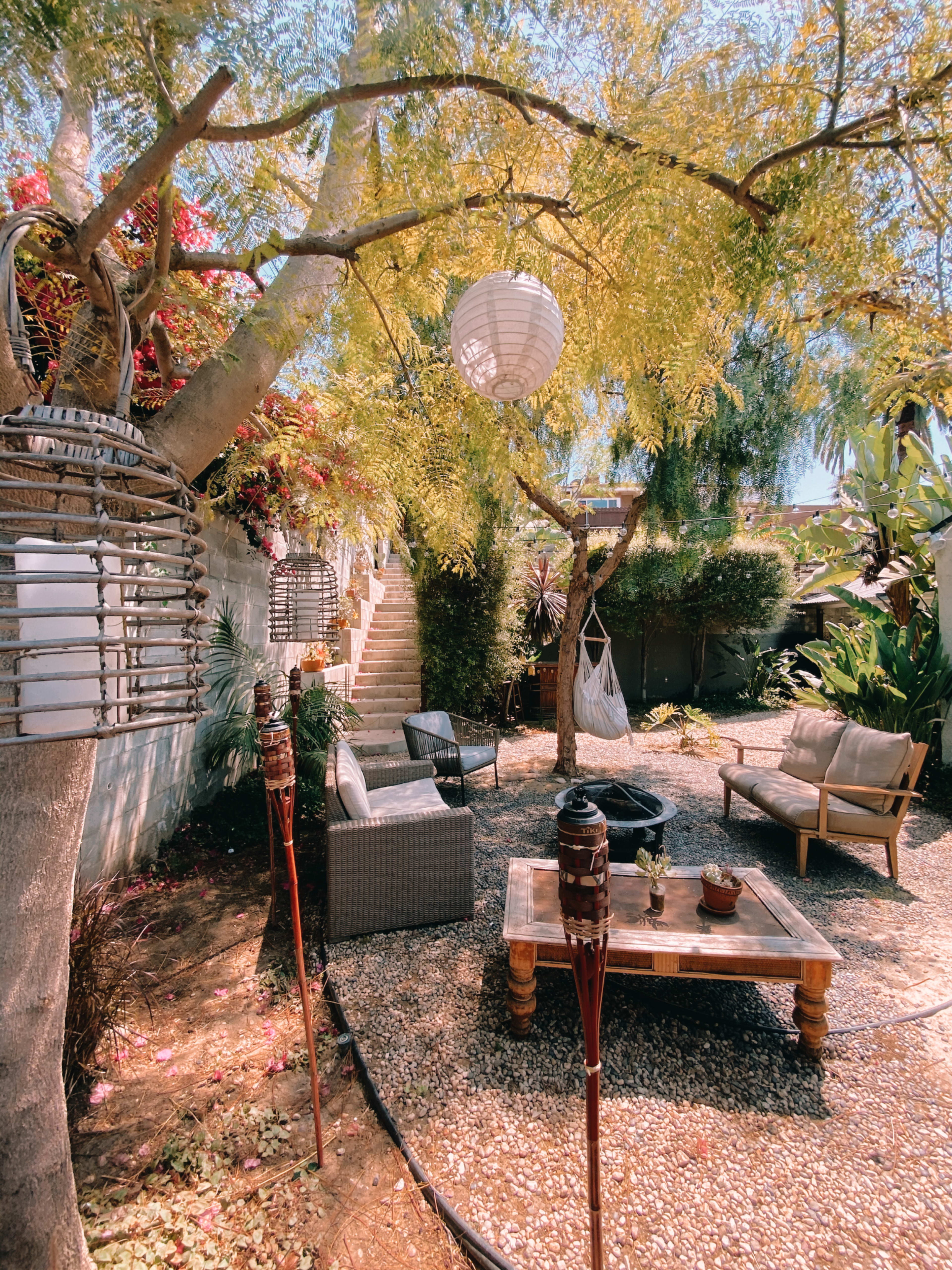 A small outdoor garden features a seating area with a table and chairs, surrounded by greenery and decorative lanterns.
