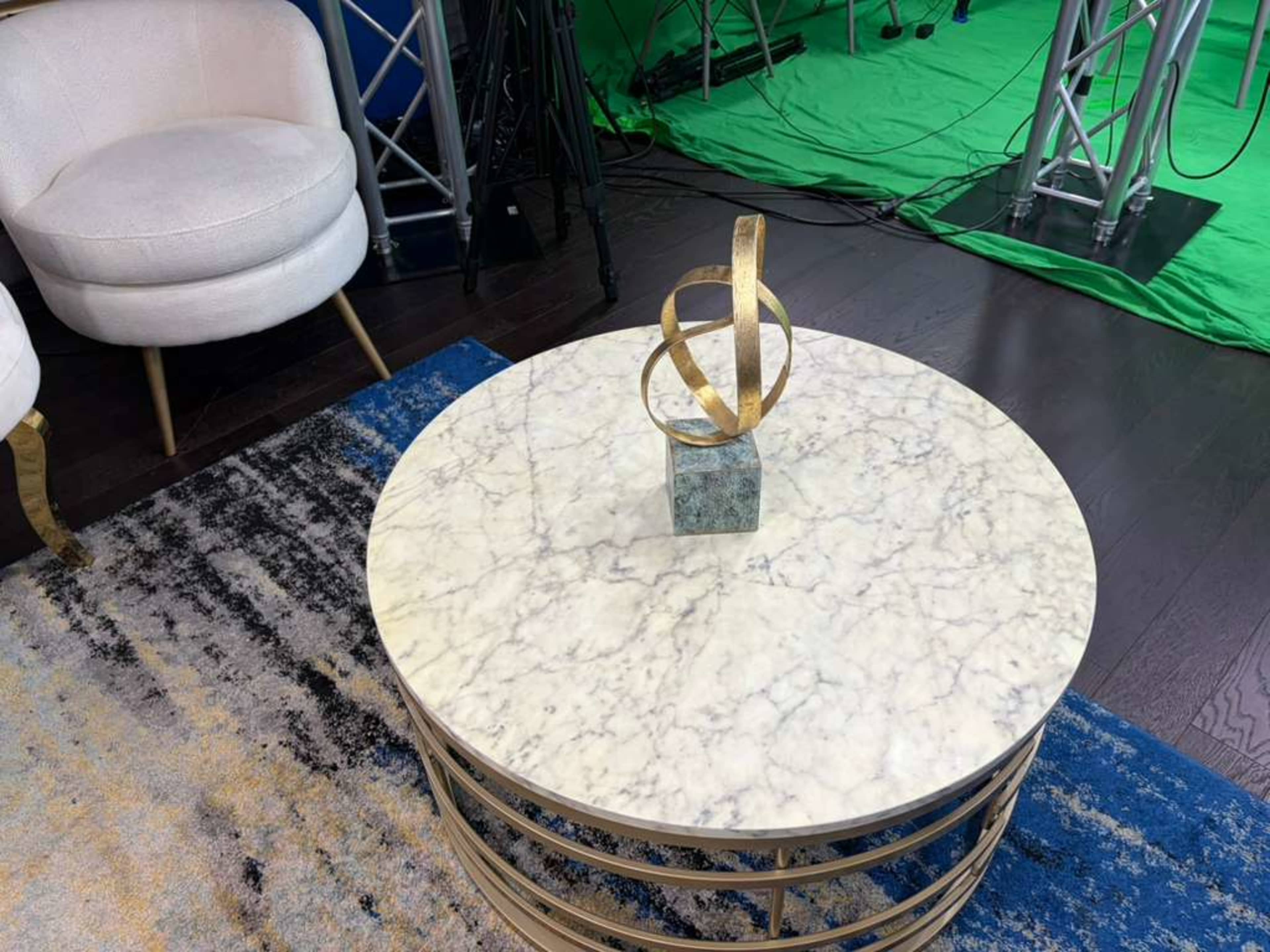 The image shows a coffee table with a marble top, a decorative sculpture, and is situated in a room with a green backdrop and studio equipment.