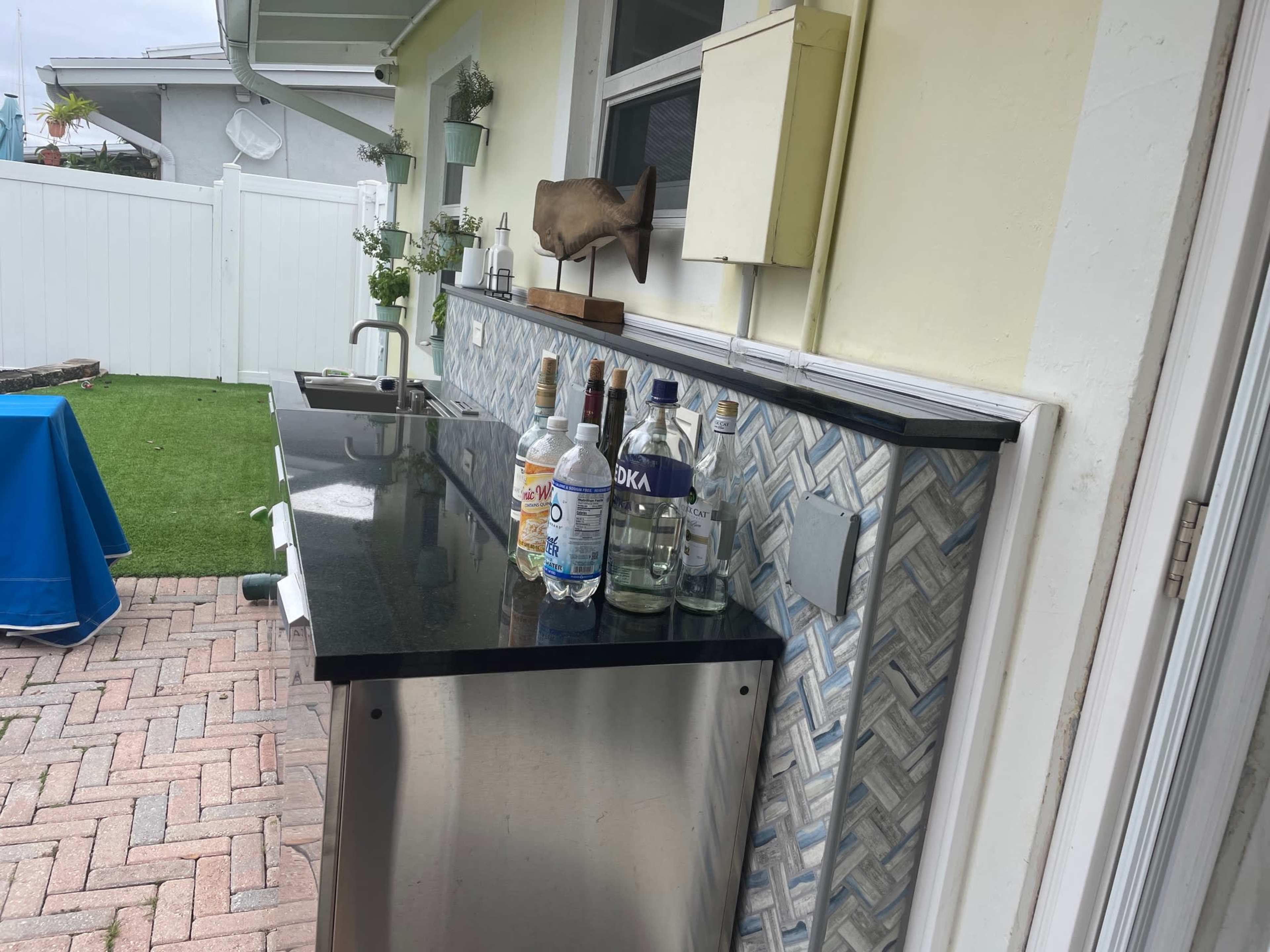 The image shows an outdoor bar area with a black countertop, a stainless steel base, and several bottles of alcohol lined up against a patterned backsplash.