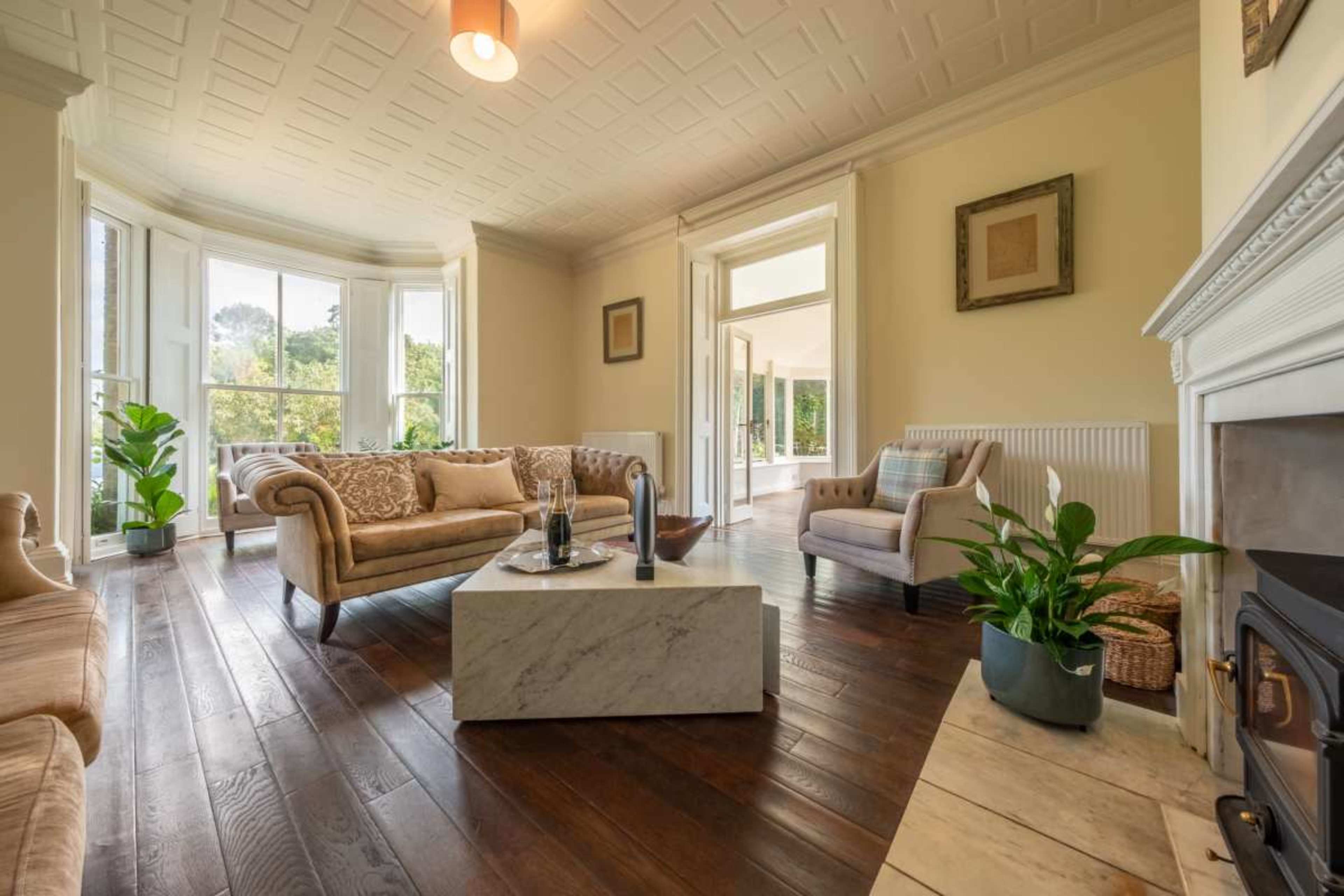 The image shows a spacious living room with large windows, a marble coffee table, and a fireplace, featuring light-colored walls and wooden flooring.