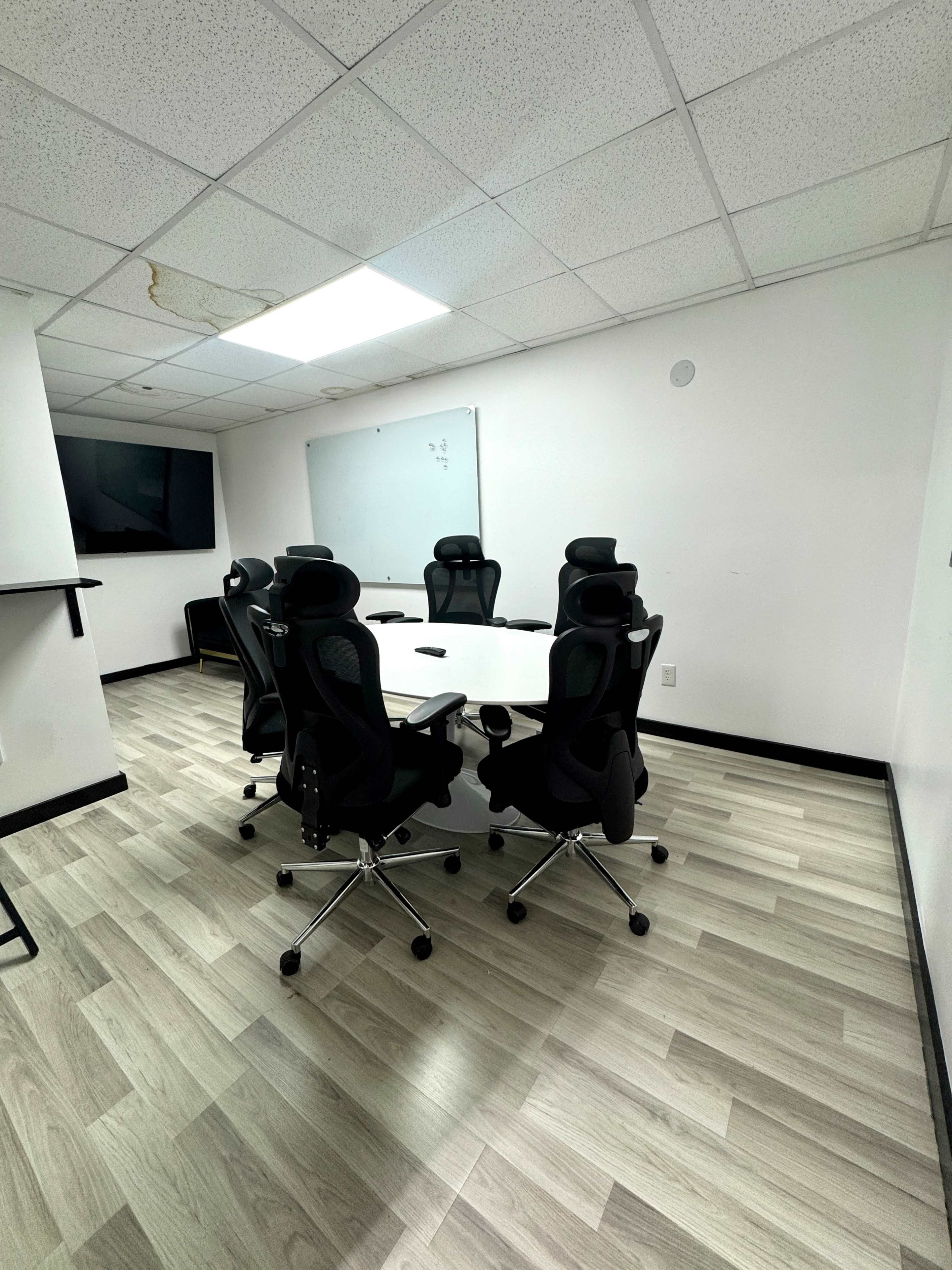 The image shows a conference room with a round table surrounded by six ergonomic chairs, a large TV on one wall, and a whiteboard on another.