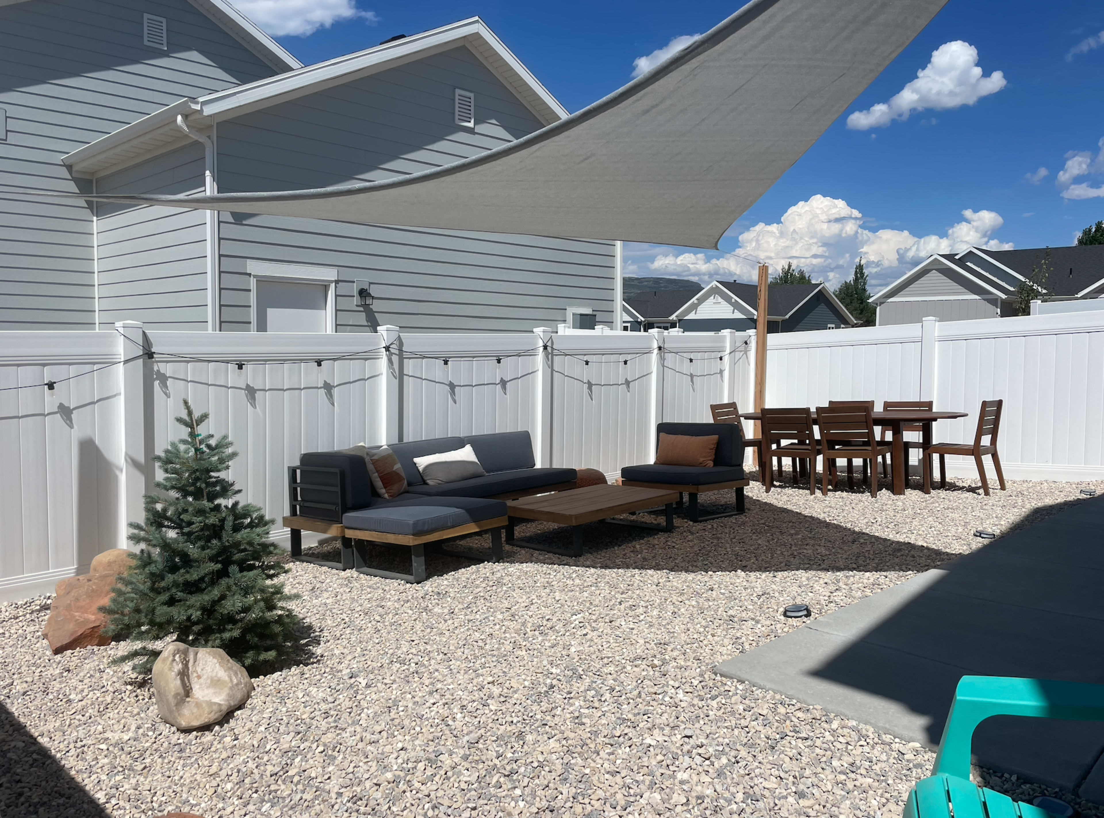 A patio area featuring a seating arrangement with a low table, a dining set, and a small pine tree, all beneath a large shade sail.