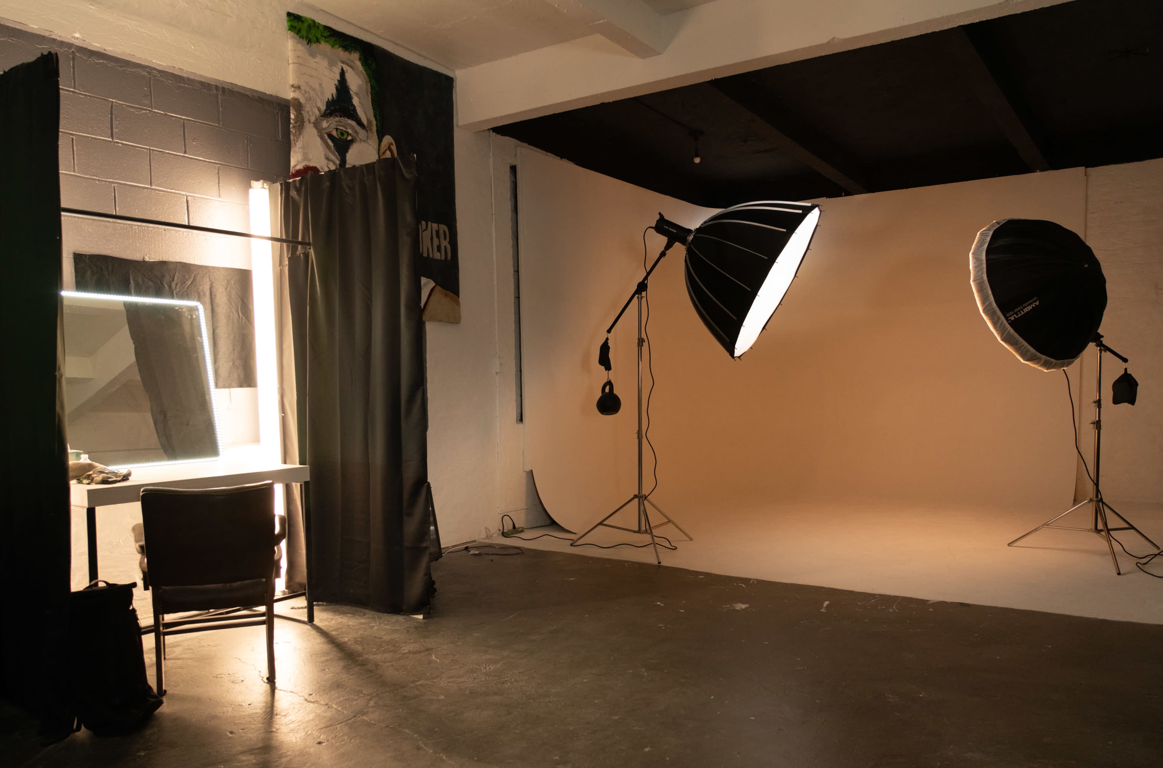 The image shows a photography studio featuring a large backdrop, two bright softbox lights, a mirror with a lighted frame, and a chair beside a black curtain.