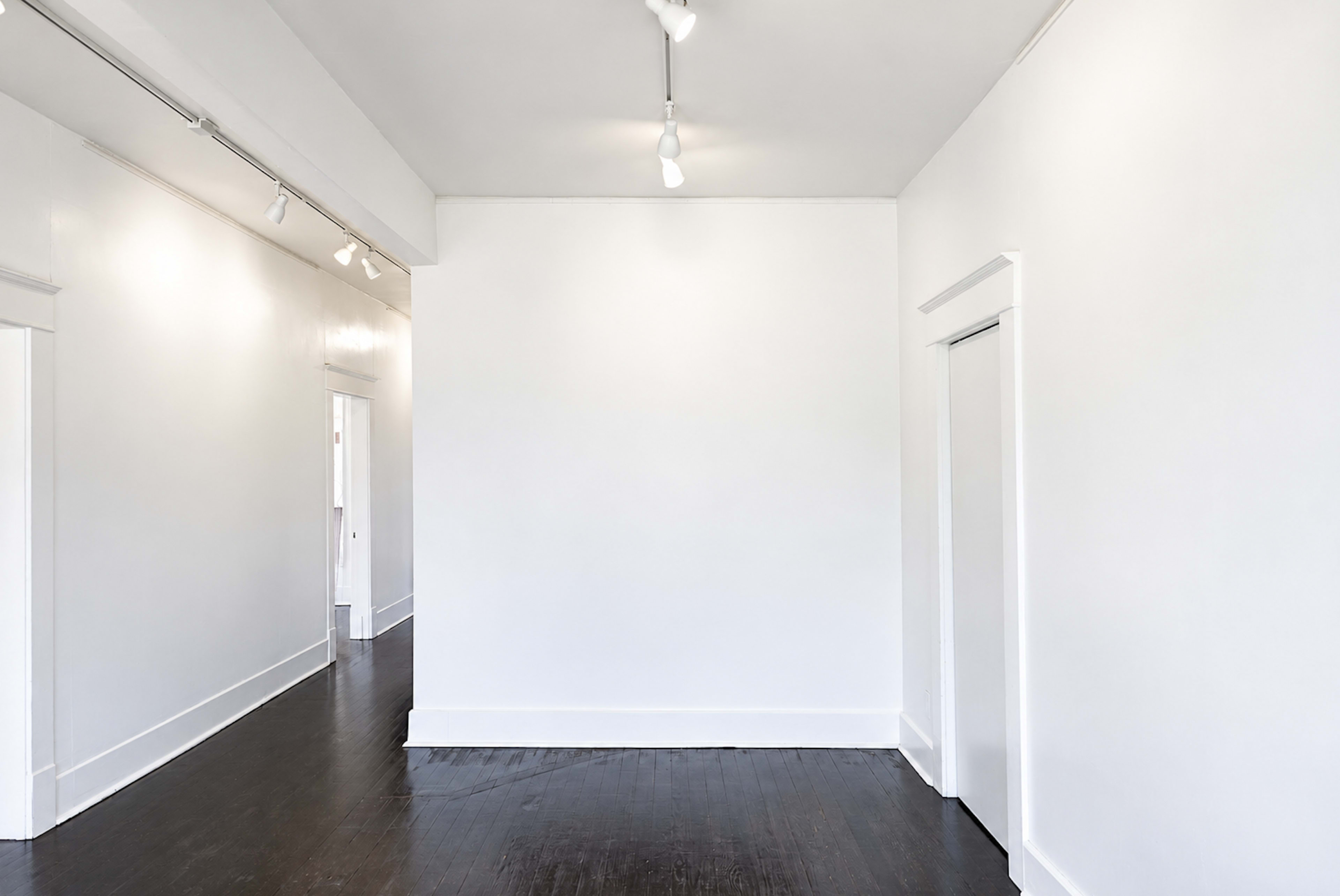 The image shows a bright, empty room with white walls and dark wooden flooring, and an open doorway leading to another space.