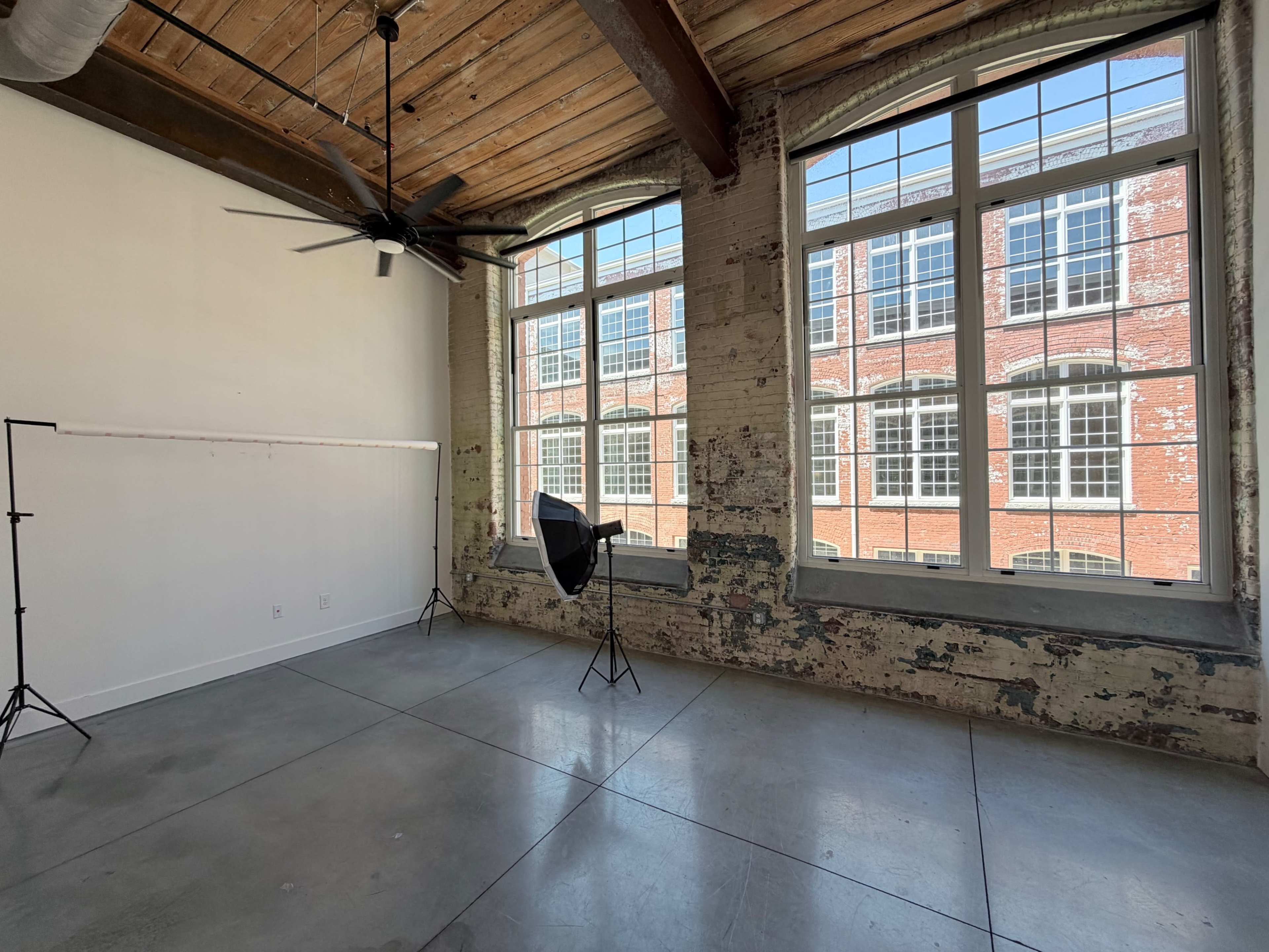 The image shows an industrial-style room with large windows, exposed brick walls, and concrete flooring.