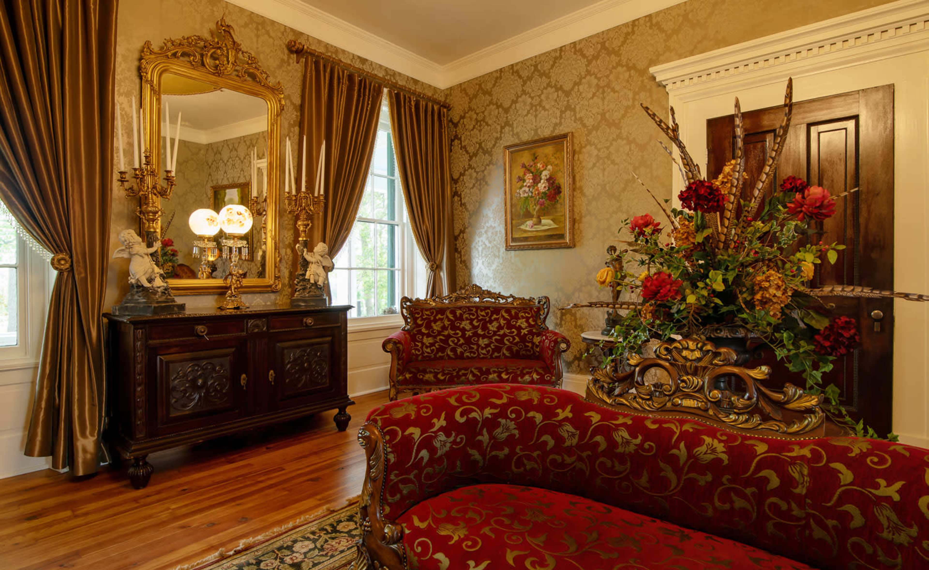 The room features ornate furniture, a large gilded mirror, and a floral arrangement, set against richly decorated wallpaper and elegant curtains.