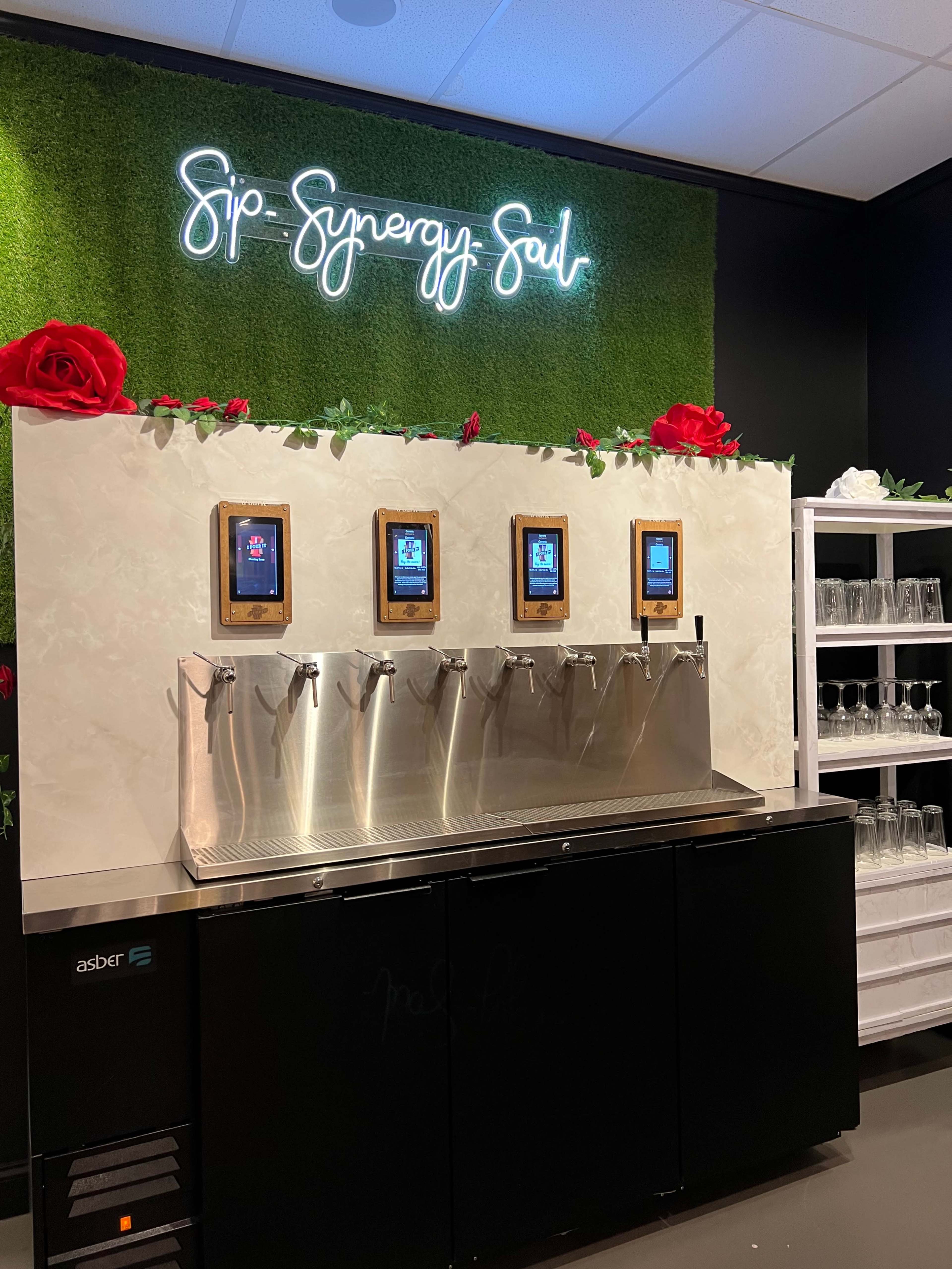 A beverage station with four dispensers, a floral-themed backdrop, and a neon sign that reads "Sip Synergy Soul."
