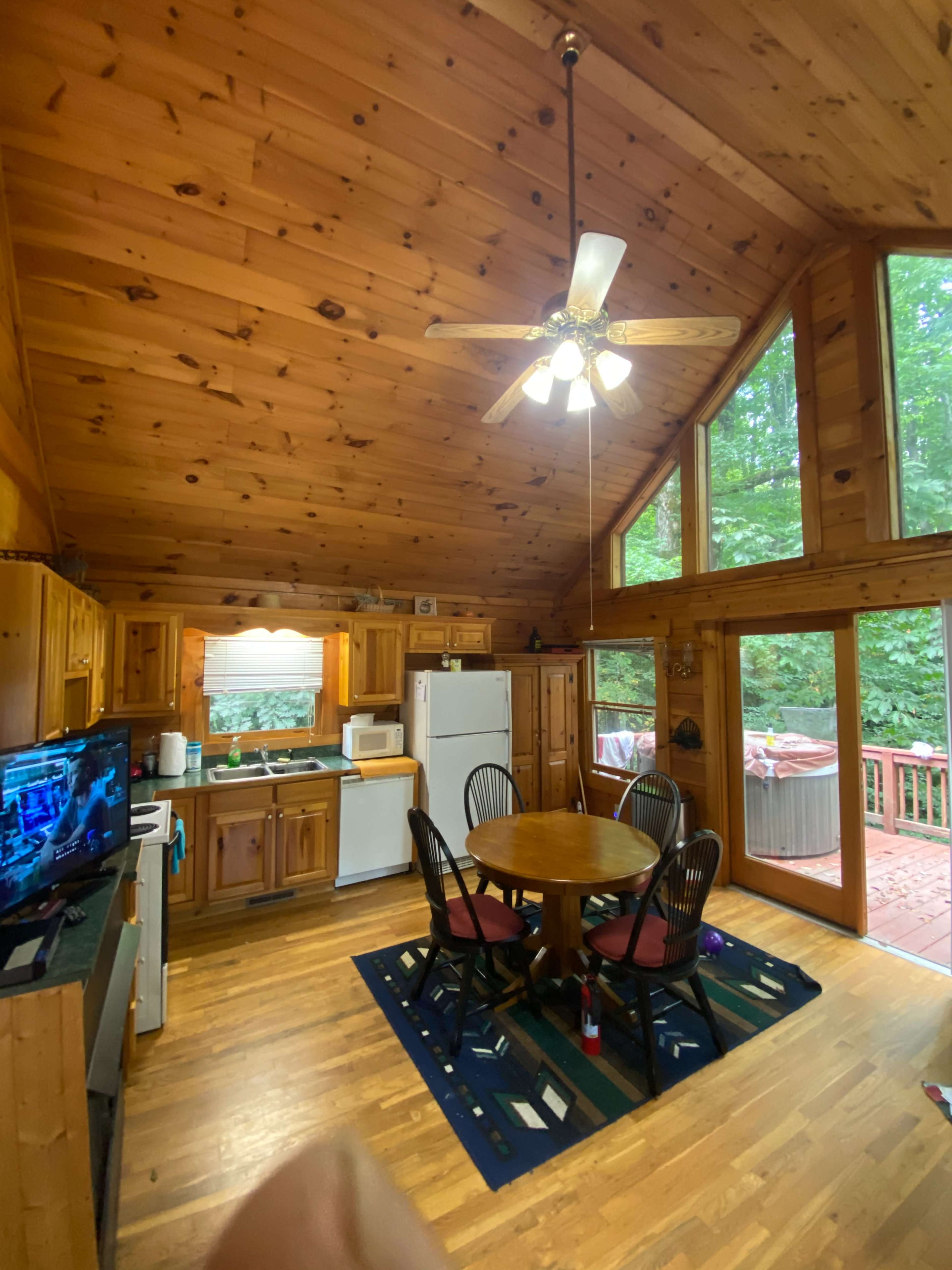 The image shows a wooden cabin kitchen with a circular dining table, four chairs, and large windows overlooking a deck.