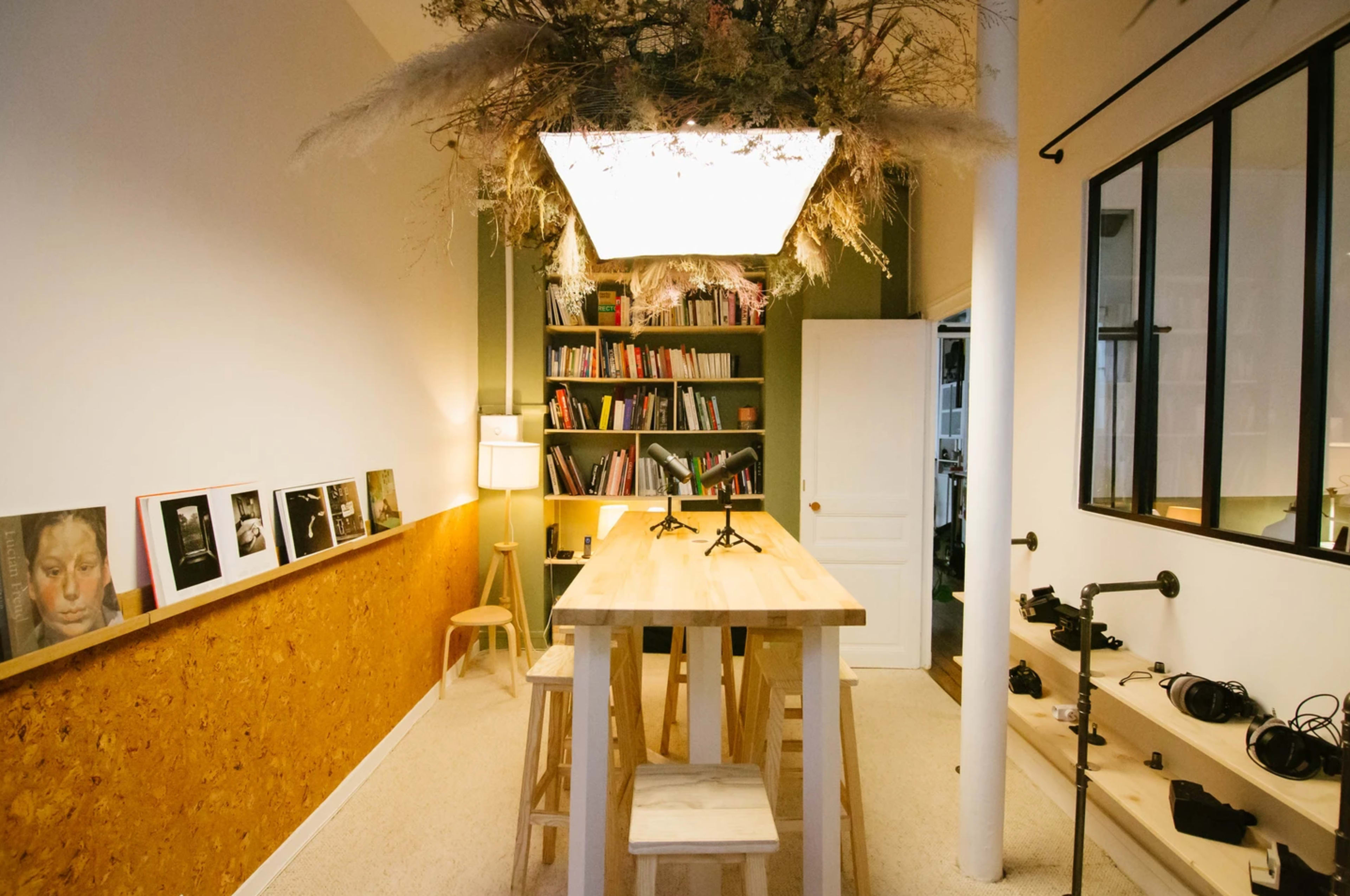 A spacious room features a long wooden table with stools, surrounded by bookshelves, photography, and a large overhead light decorated with plants.