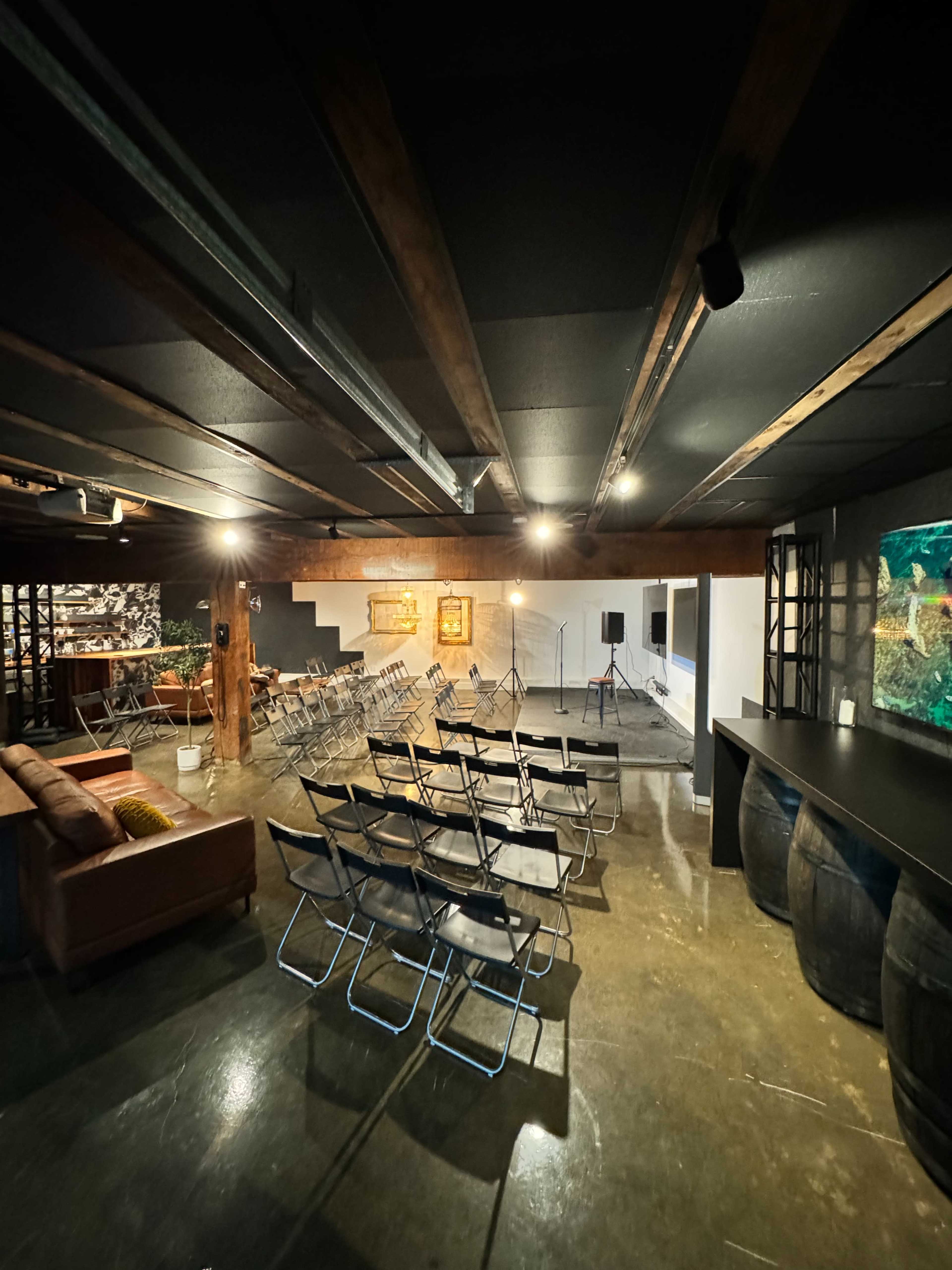 A dimly lit event space featuring rows of folding chairs, a seating area with a couch, and a bar made from barrels.