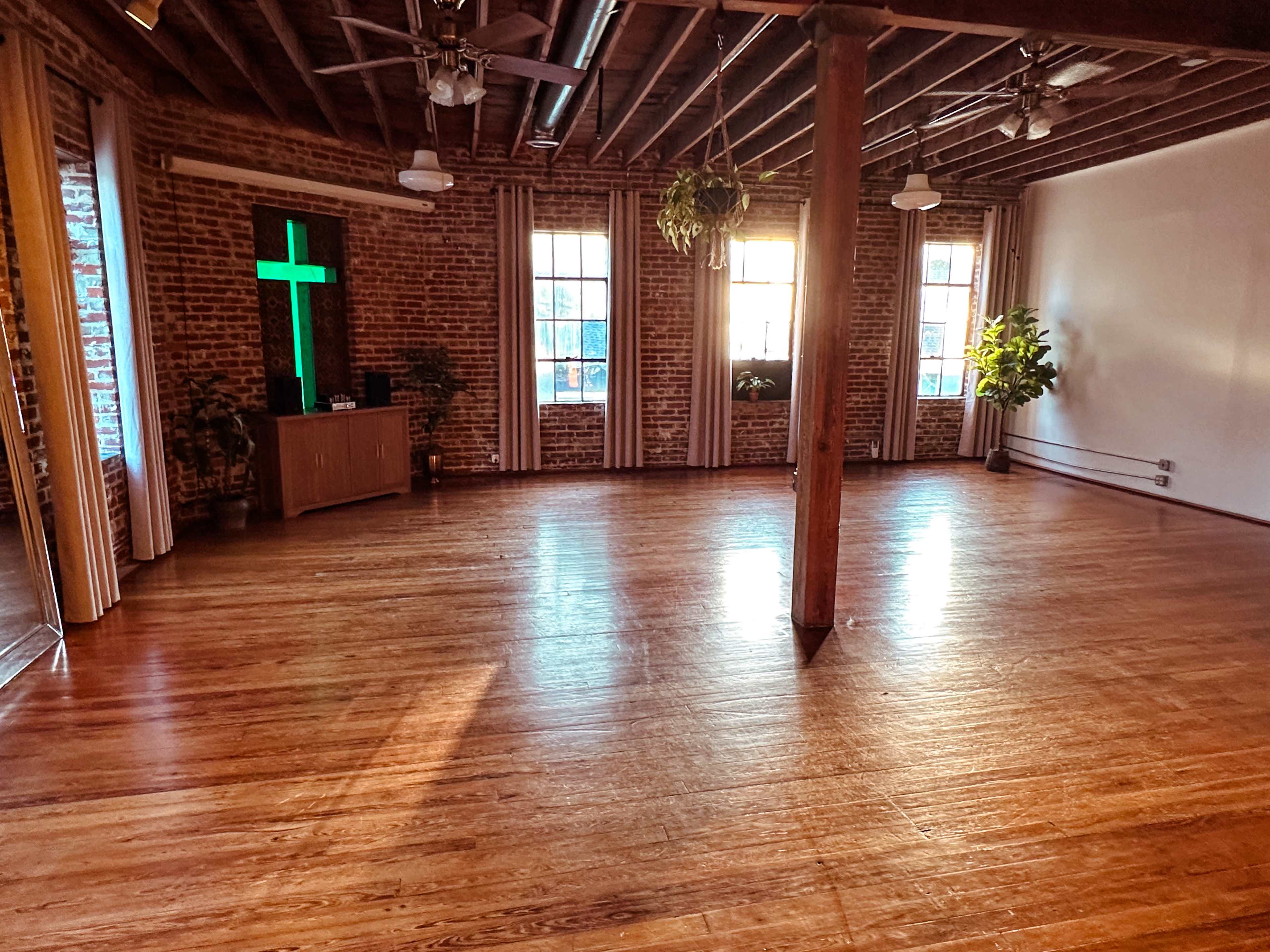 The image shows a spacious, wooden-floored room with a brick wall, a green illuminated cross, and several plants near the windows.