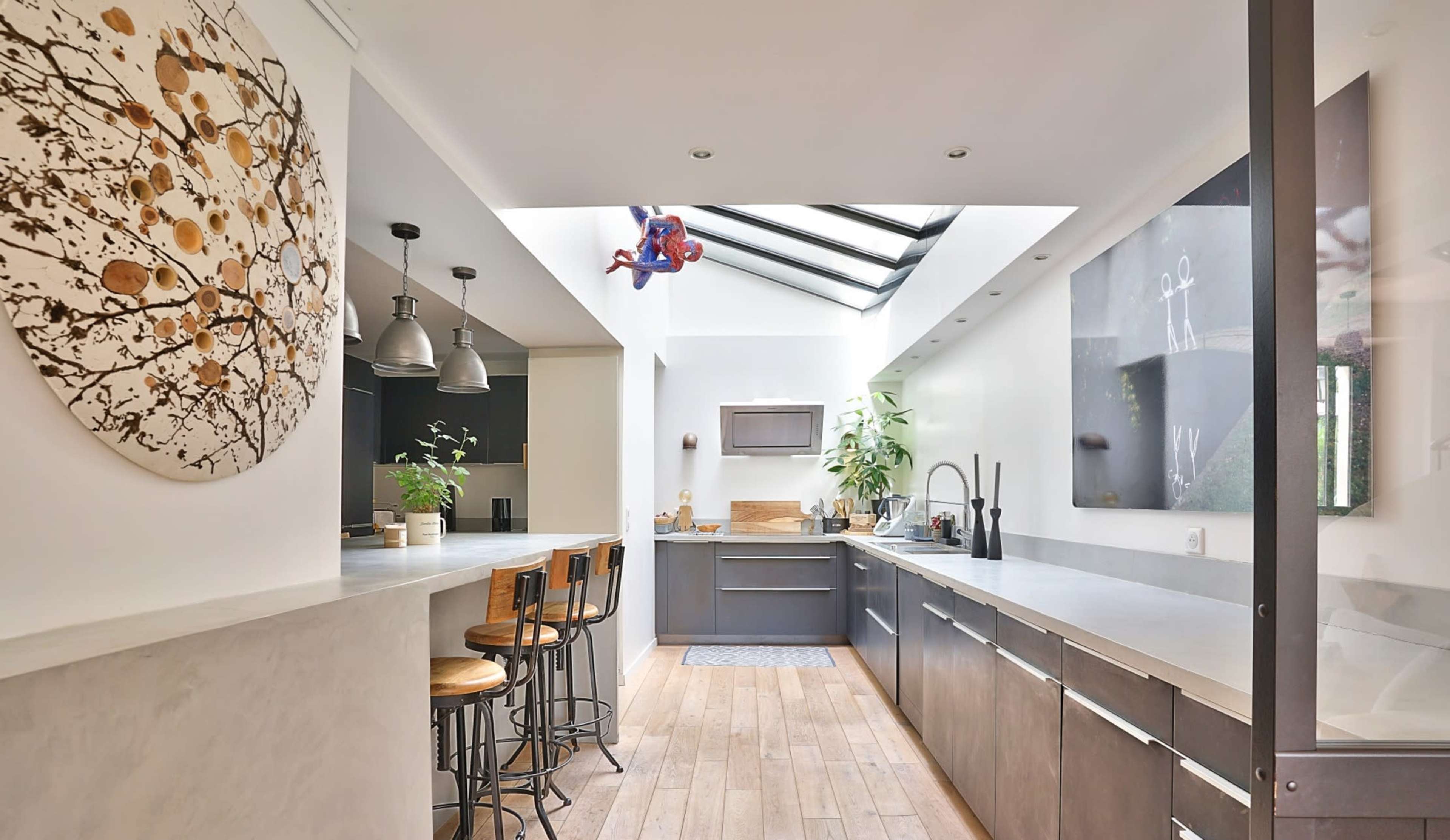 A modern kitchen with wooden flooring, gray cabinetry, and a skylight, featuring a Spider-Man figure suspended from the ceiling.