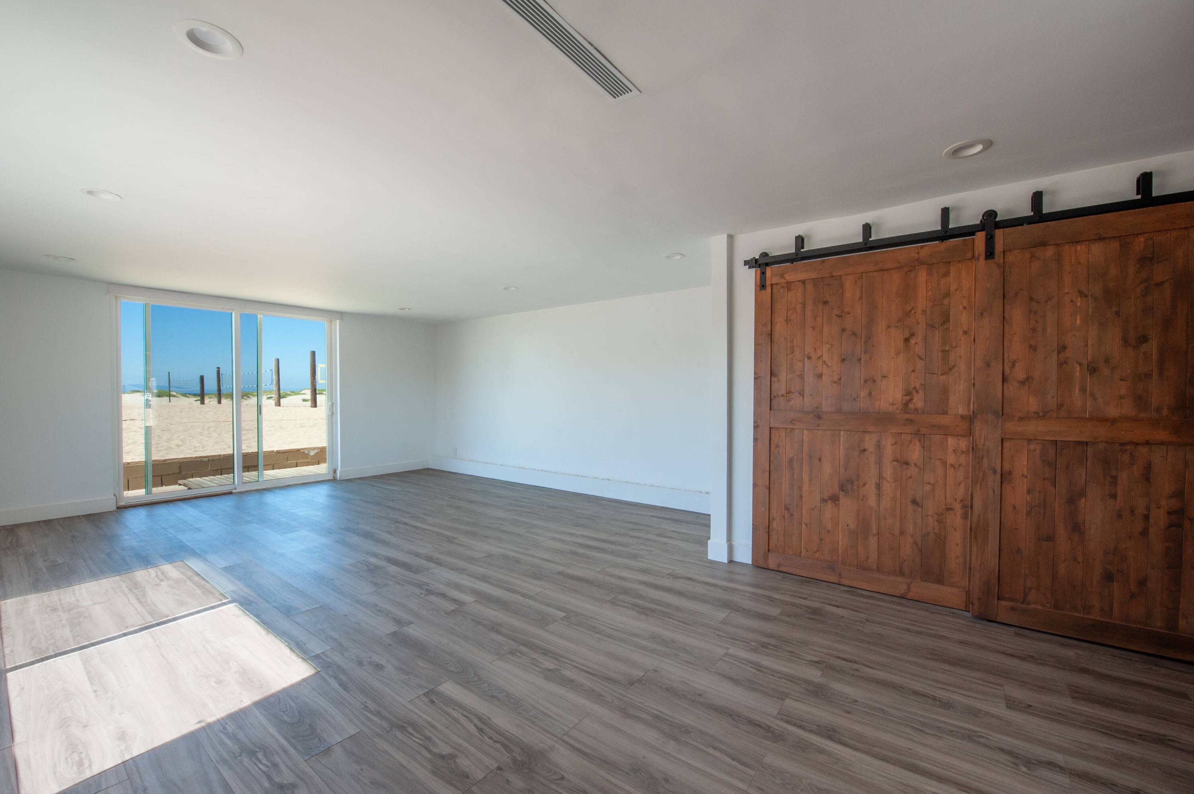 The image shows an empty room with wooden sliding doors and large windows facing a sandy outdoor area.