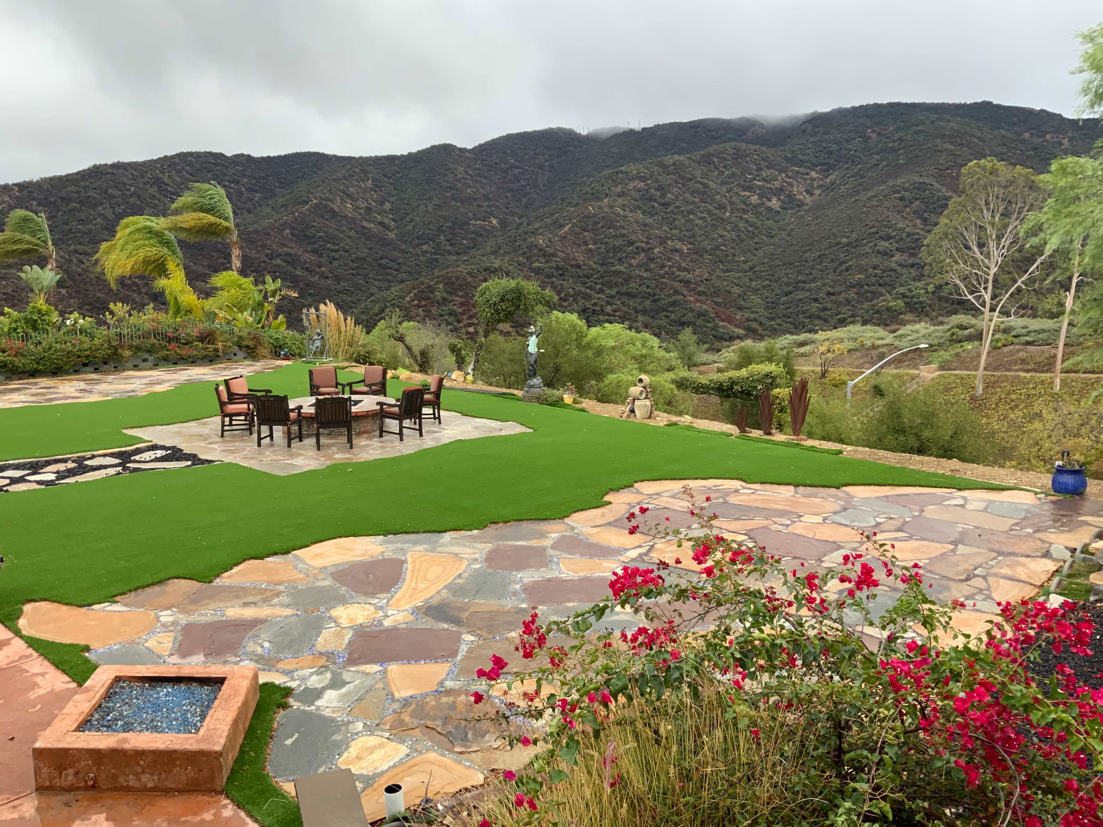 A landscaped outdoor area with a stone patio, a seating arrangement around a table, and a mountainous backdrop.