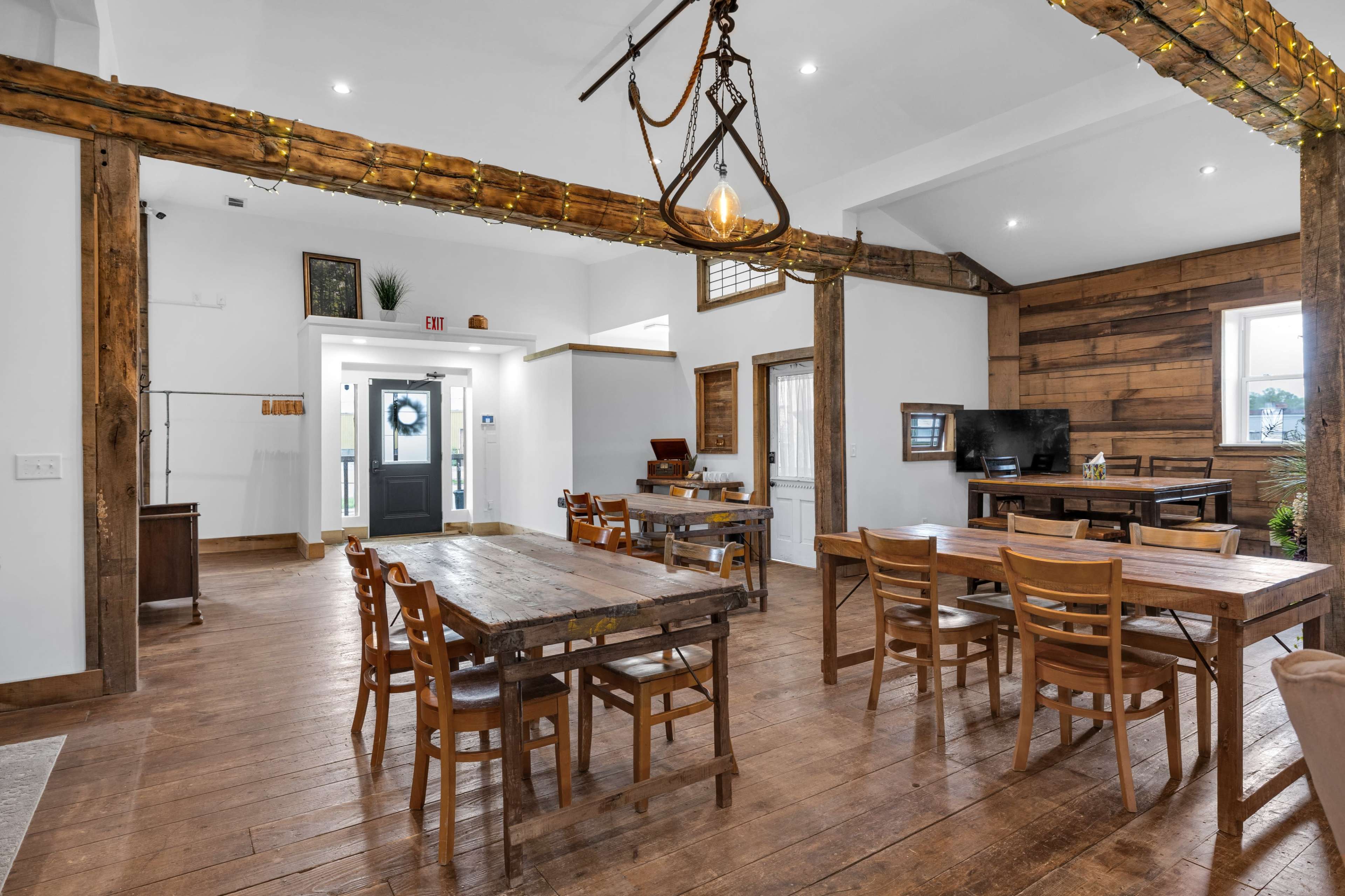 The image shows a spacious dining area with wooden tables and chairs, exposed beams, and a front entrance featuring a decorative wreath.
