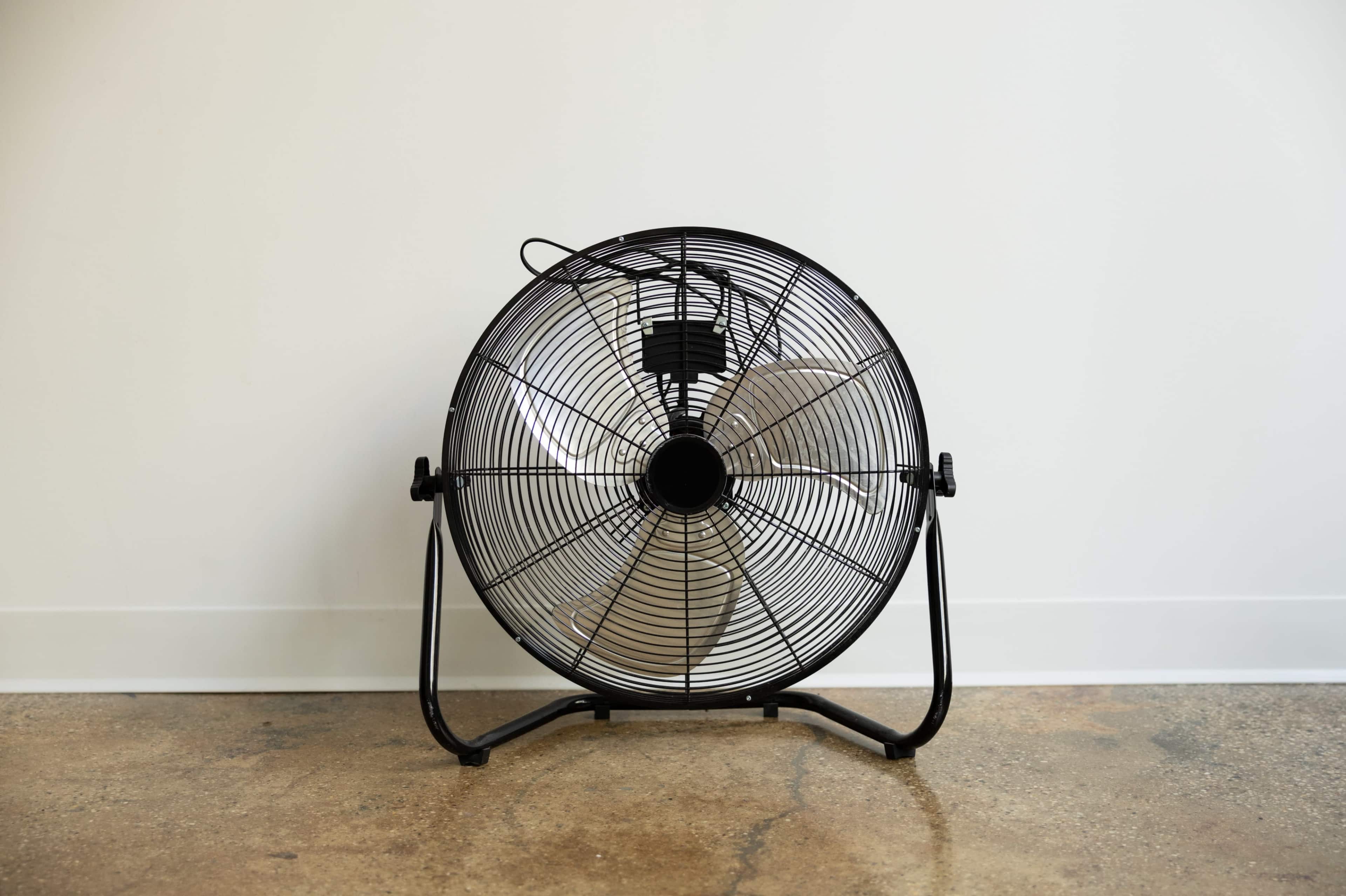 A black metal stand fan is positioned against a plain white wall on a concrete floor.