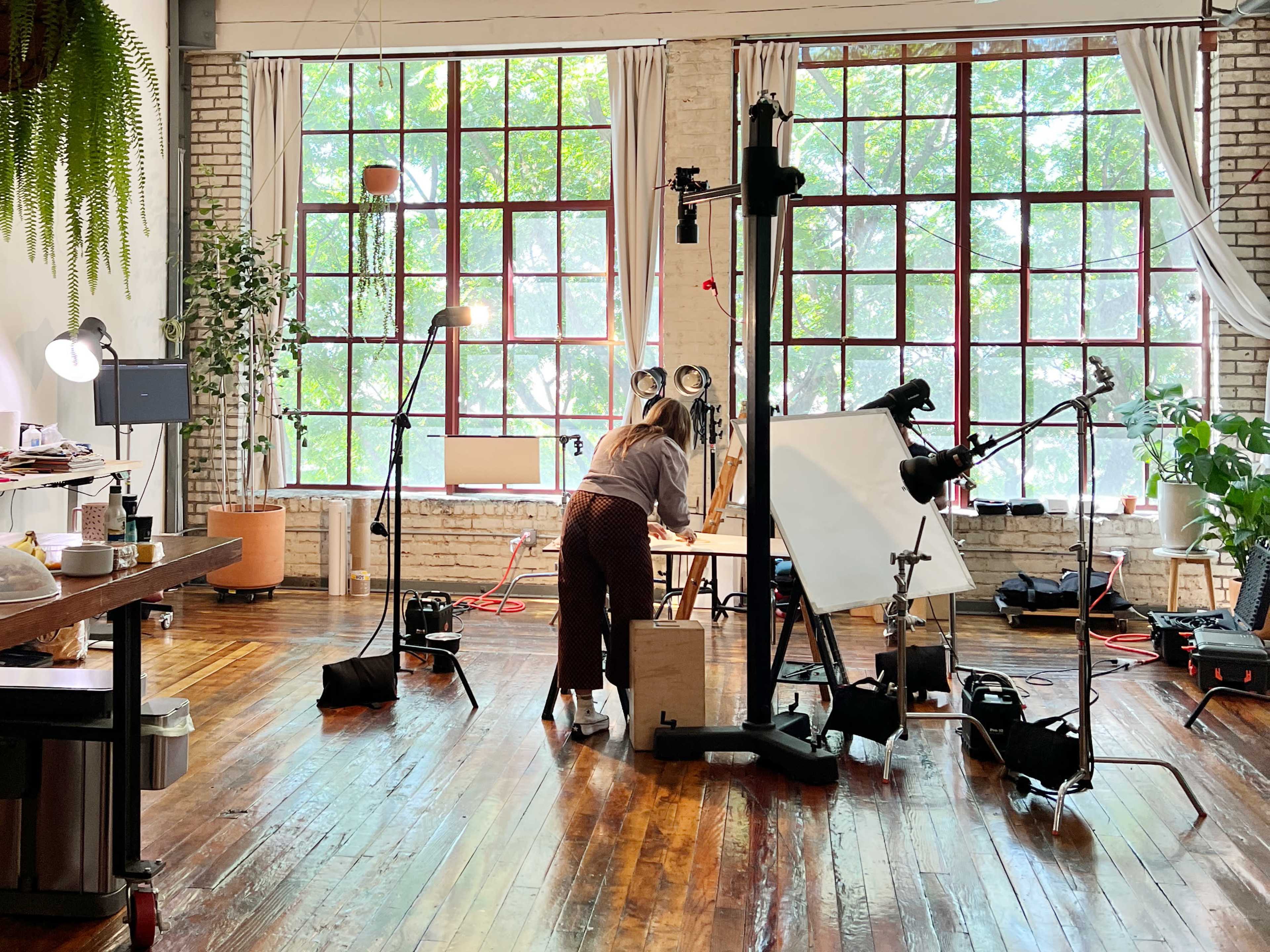 A woman works at a table in a spacious studio with large windows, surrounded by various lighting equipment and plants.
