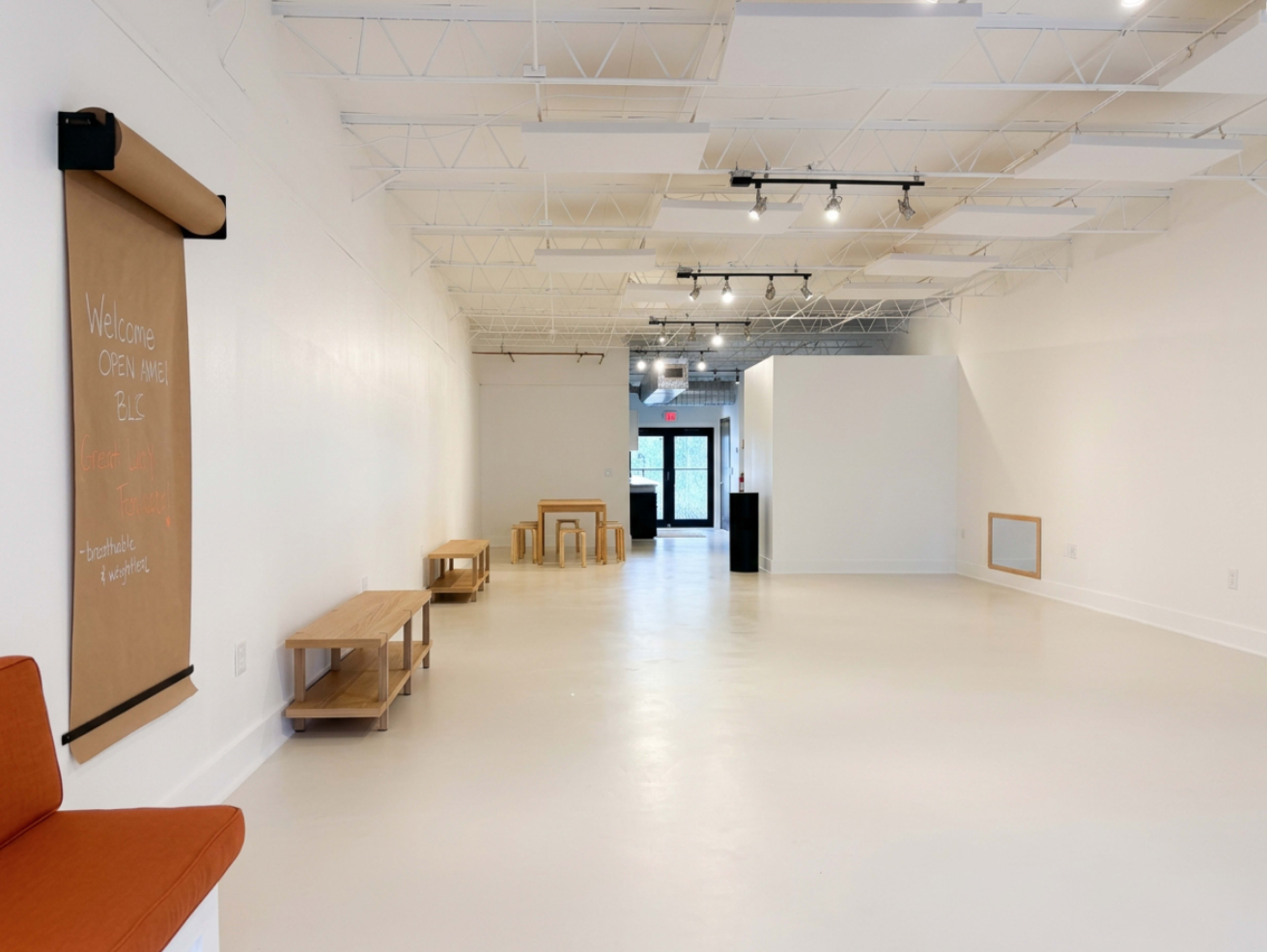 The image shows a spacious, well-lit interior featuring a few wooden benches and tables, with a large blank white wall at one end and an entrance visible through glass doors.