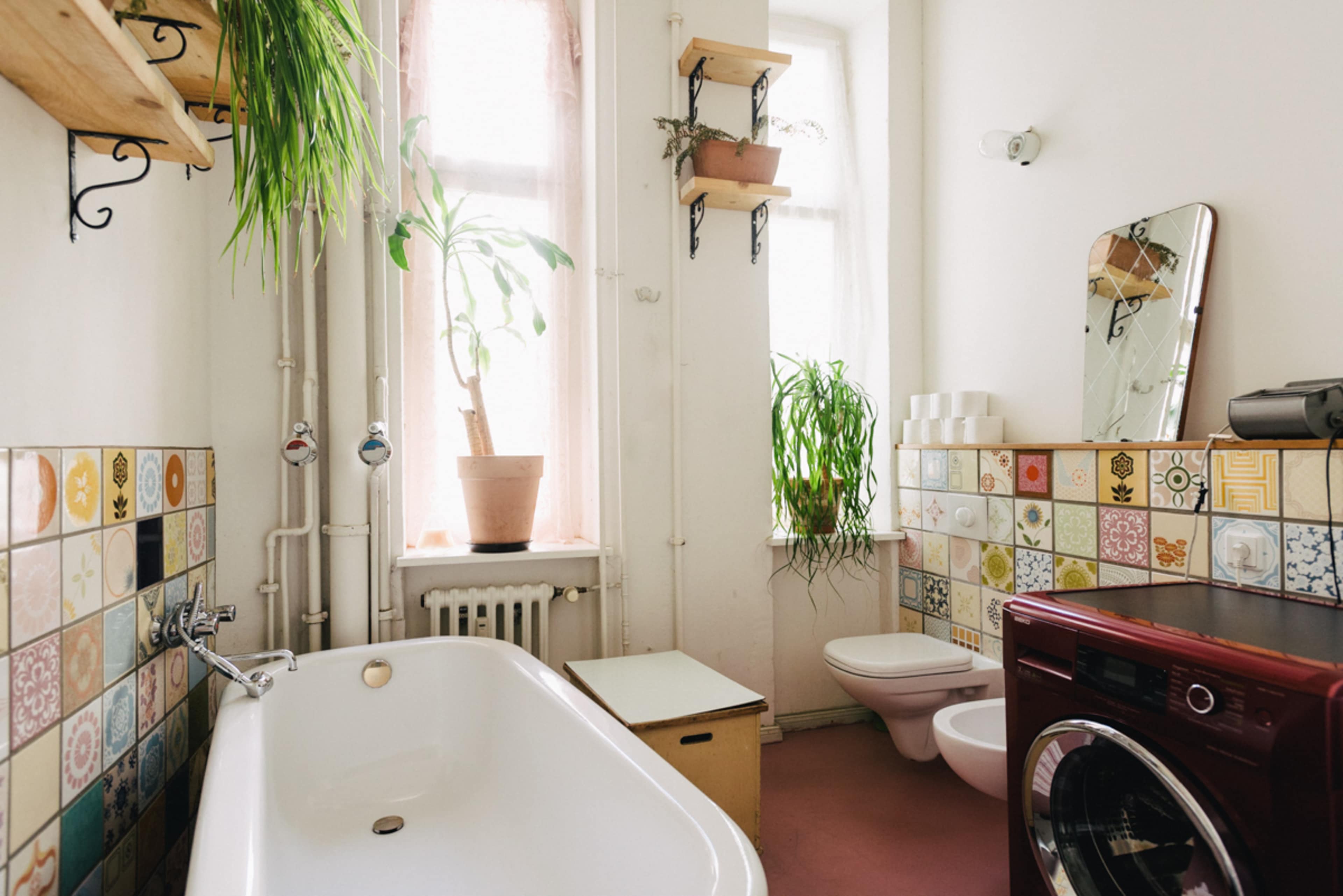 The image shows a bright bathroom with a freestanding bathtub, a toilet, a washing machine, and potted plants near a window, surrounded by colorful tiled walls.