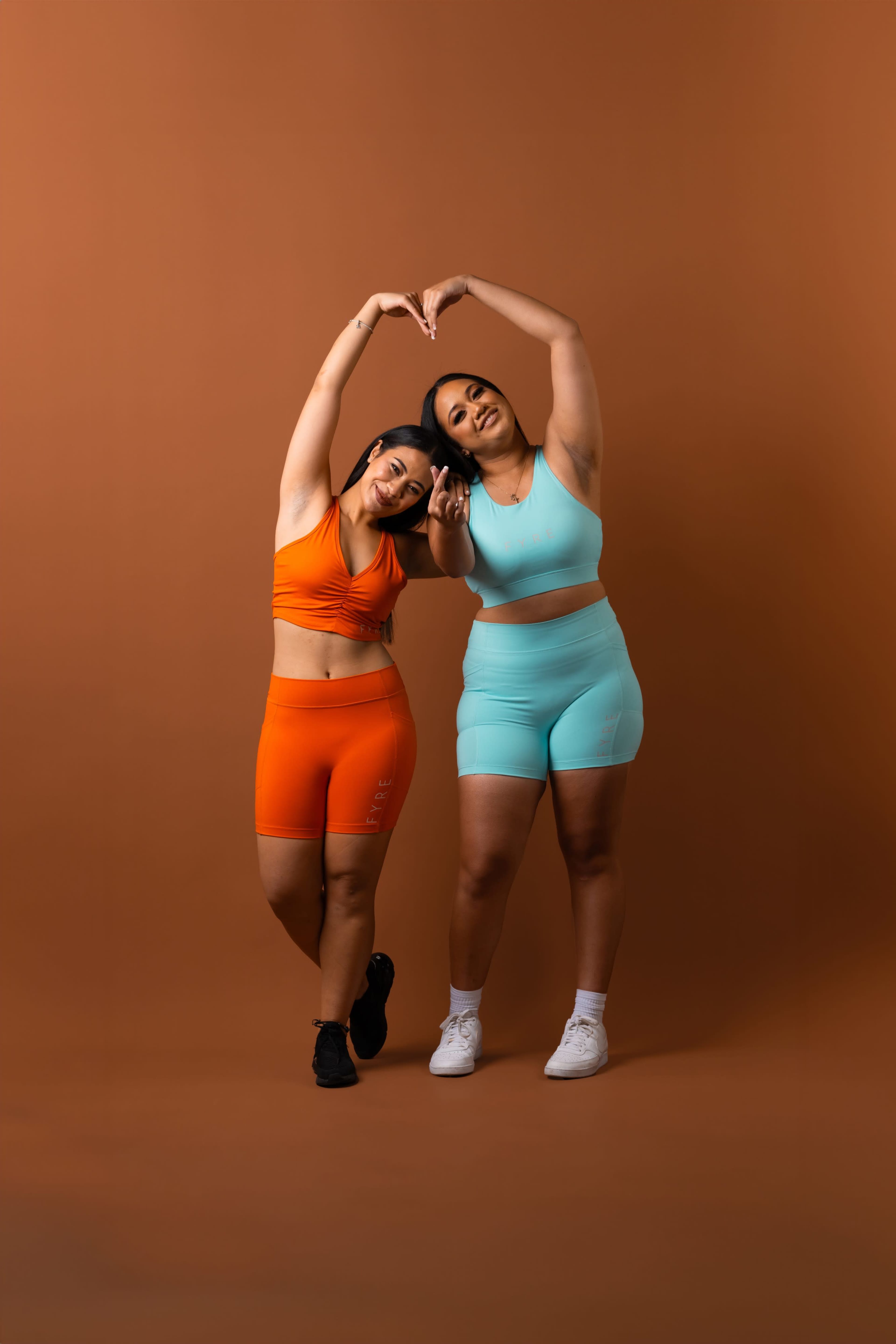 Two women in brightly colored athletic wear make a heart shape with their arms against a brown background.