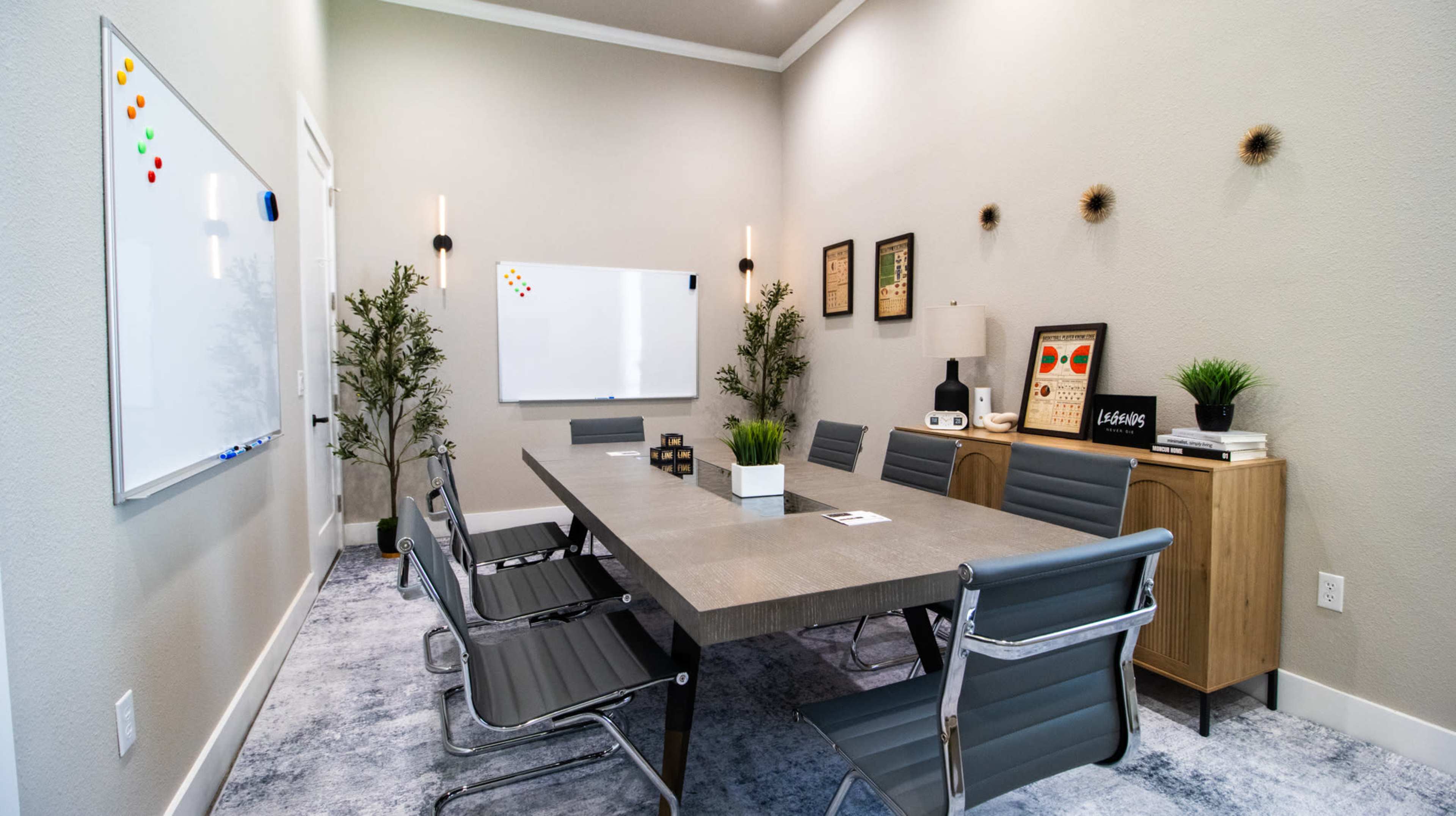 A modern conference room features a large rectangular table surrounded by six chairs, a whiteboard on one wall, and decor including plants and framed artwork.