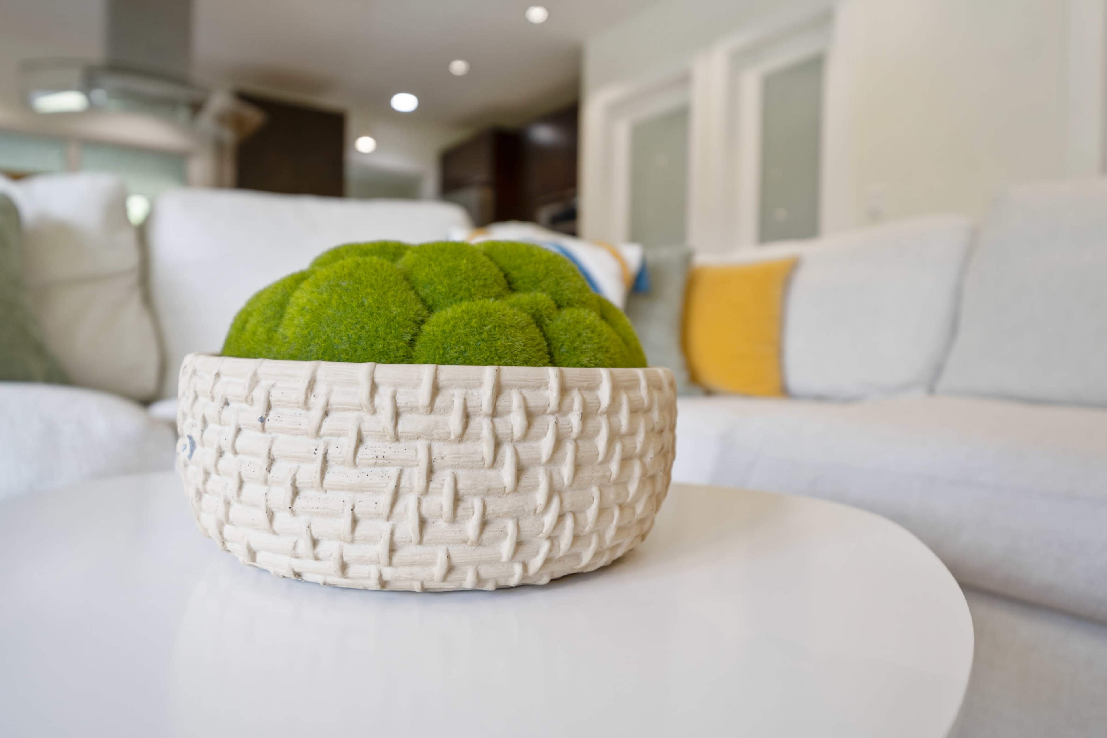 A woven basket filled with bright green decorative moss sits on a white table in a softly lit living room.