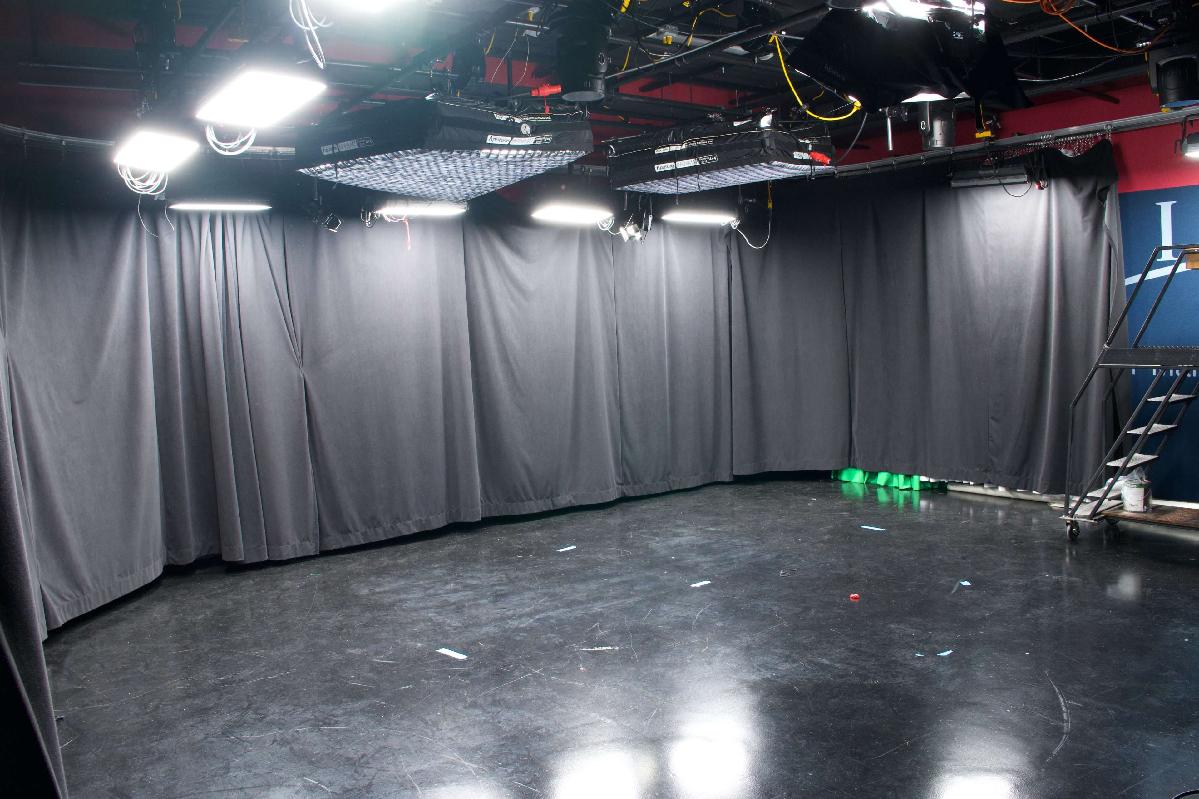 The image shows an empty studio space with black curtains, bright overhead lights, and a dark reflective floor.
