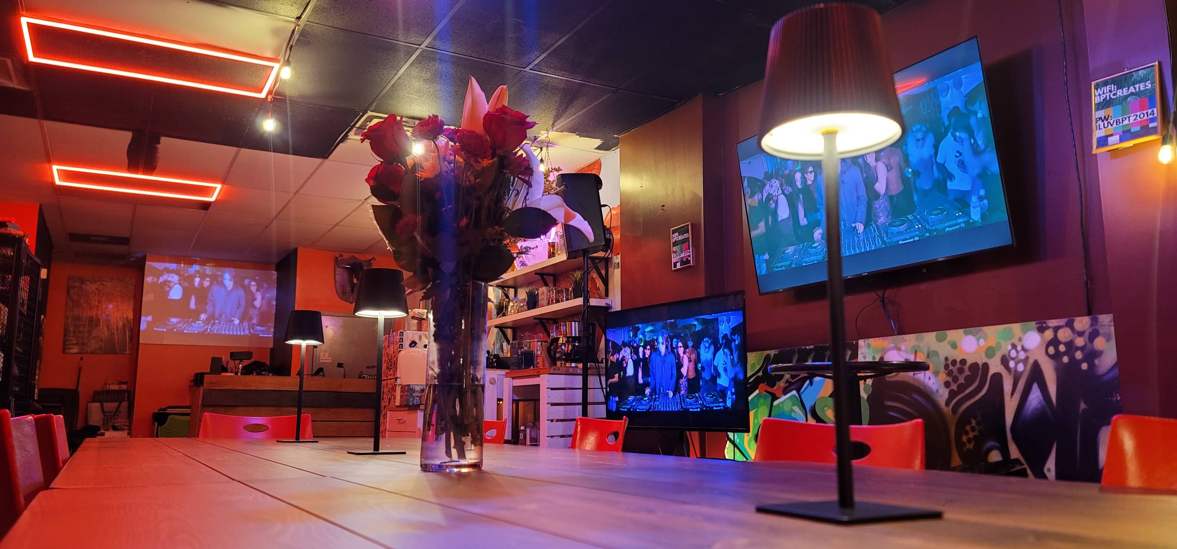 The image shows a dimly lit room with a long wooden table, red chairs, a vase of roses in the center, and two screens displaying images on the walls.
