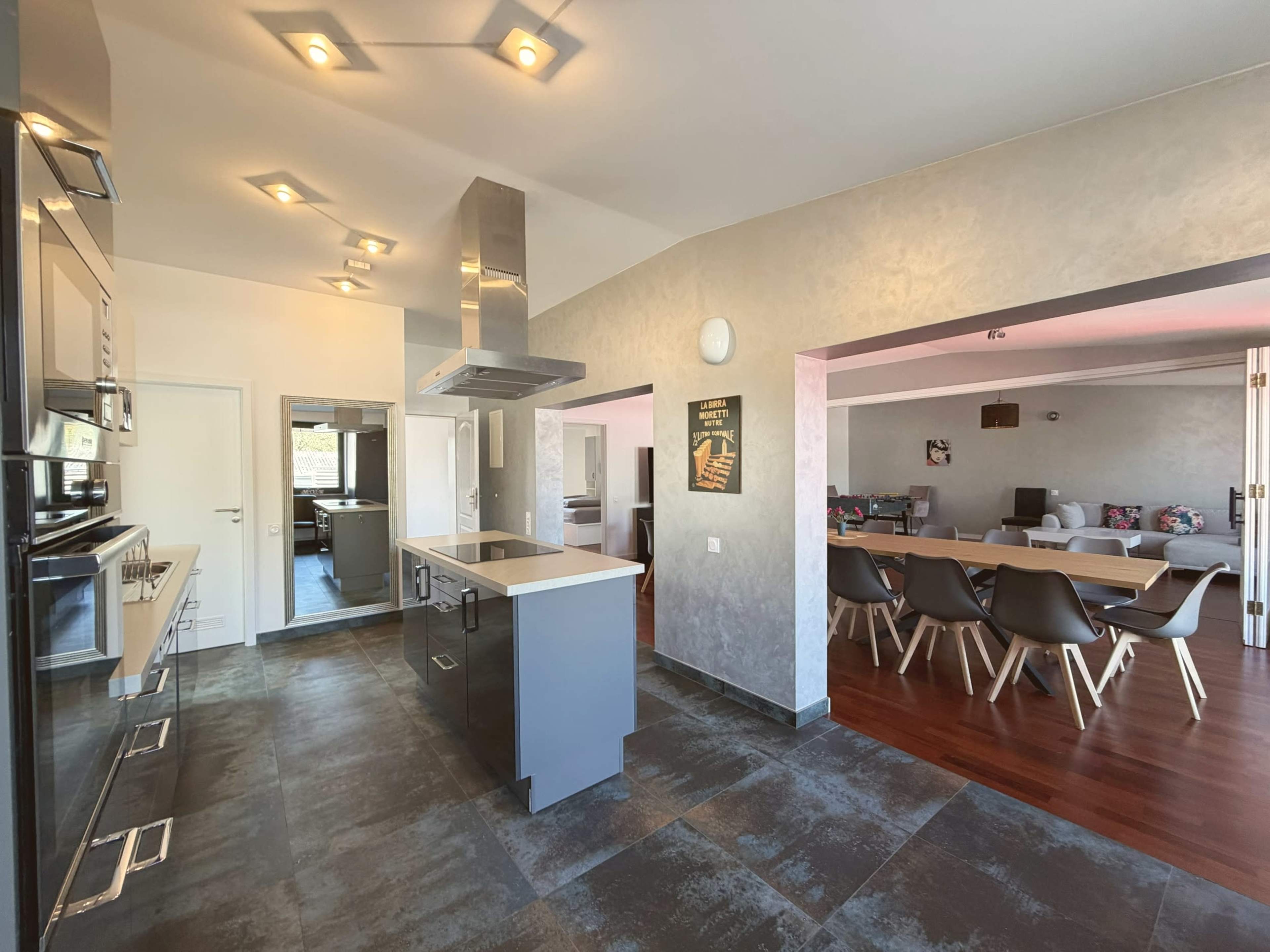 The image shows a modern kitchen with stainless steel appliances and an open layout leading to a living area furnished with a large couch and several chairs.