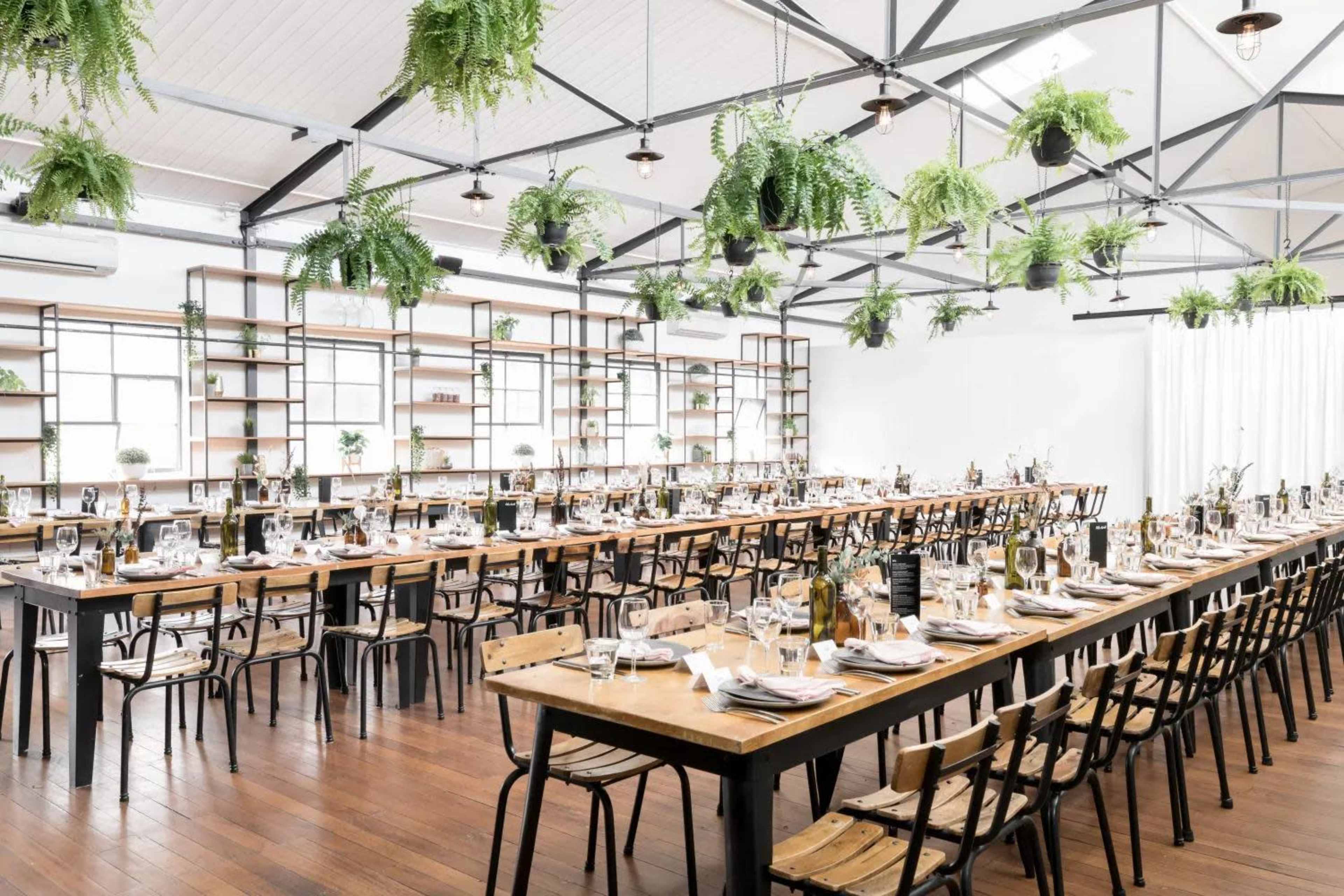 A spacious dining hall features long wooden tables arranged for a formal meal, with hanging plants suspended from the ceiling.