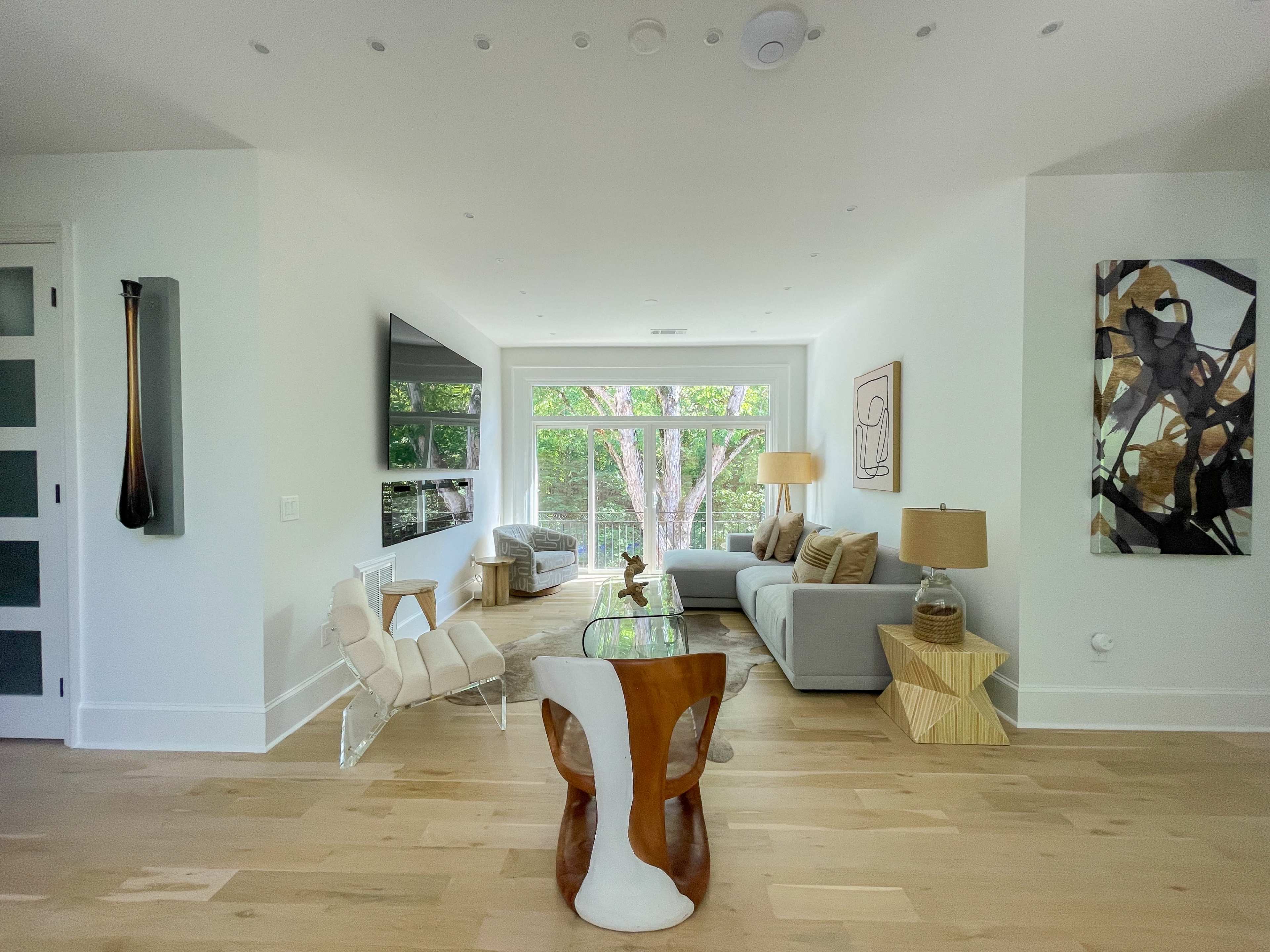 A modern living room with a gray sofa, a glass coffee table, a wooden dining table, and large windows letting in natural light.
