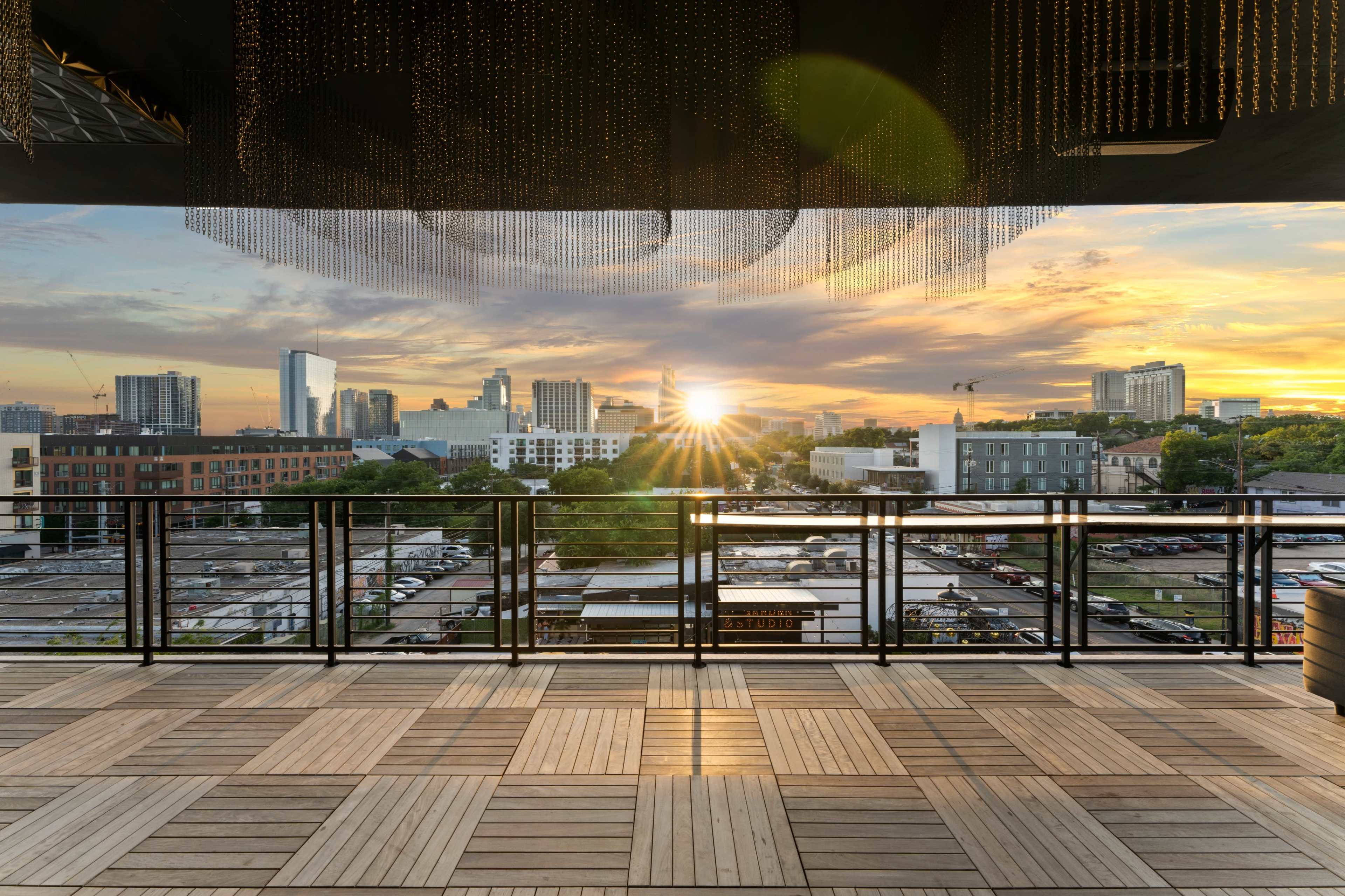 A balcony with a wooden floor overlooking a city skyline at sunset, with the sun illuminating the buildings.