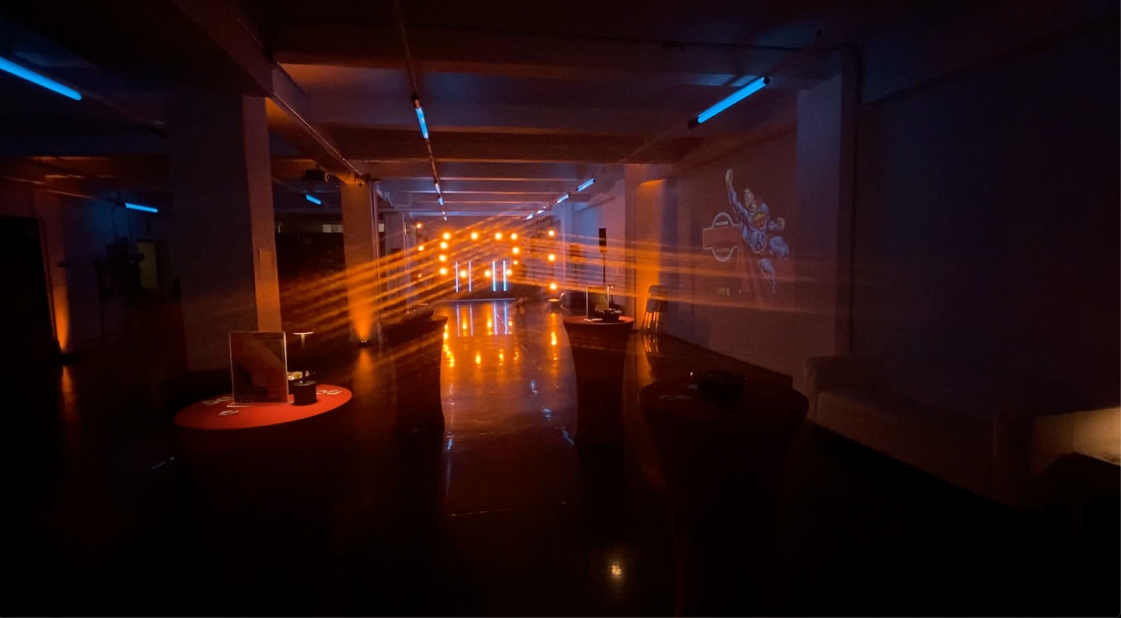 A spacious, dimly lit room with beams of light and a projection on the wall, featuring colorful lights and furniture arranged in an open layout.
