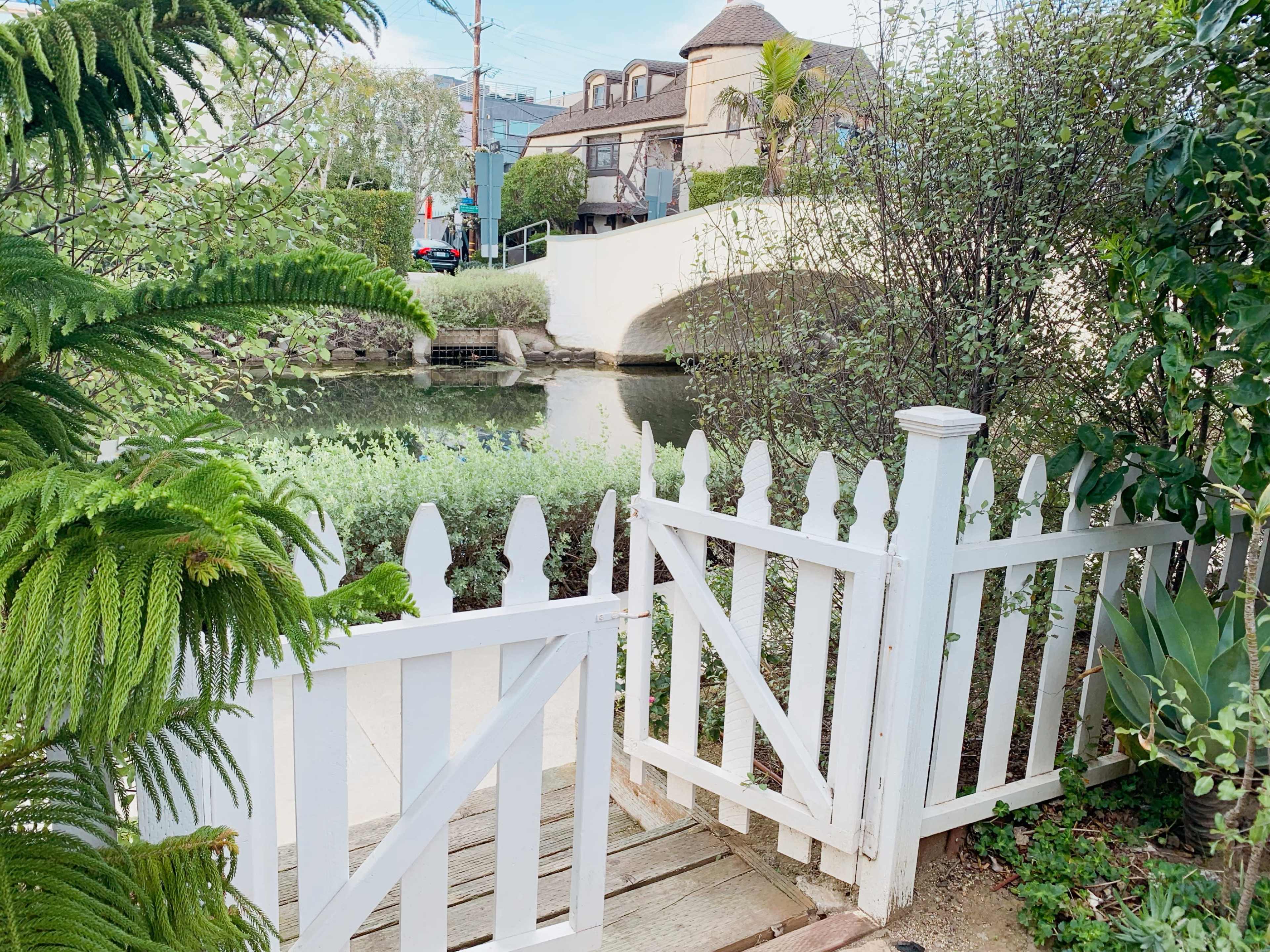 A white picket fence gate opens to a path leading to a canal with a bridge in the background.