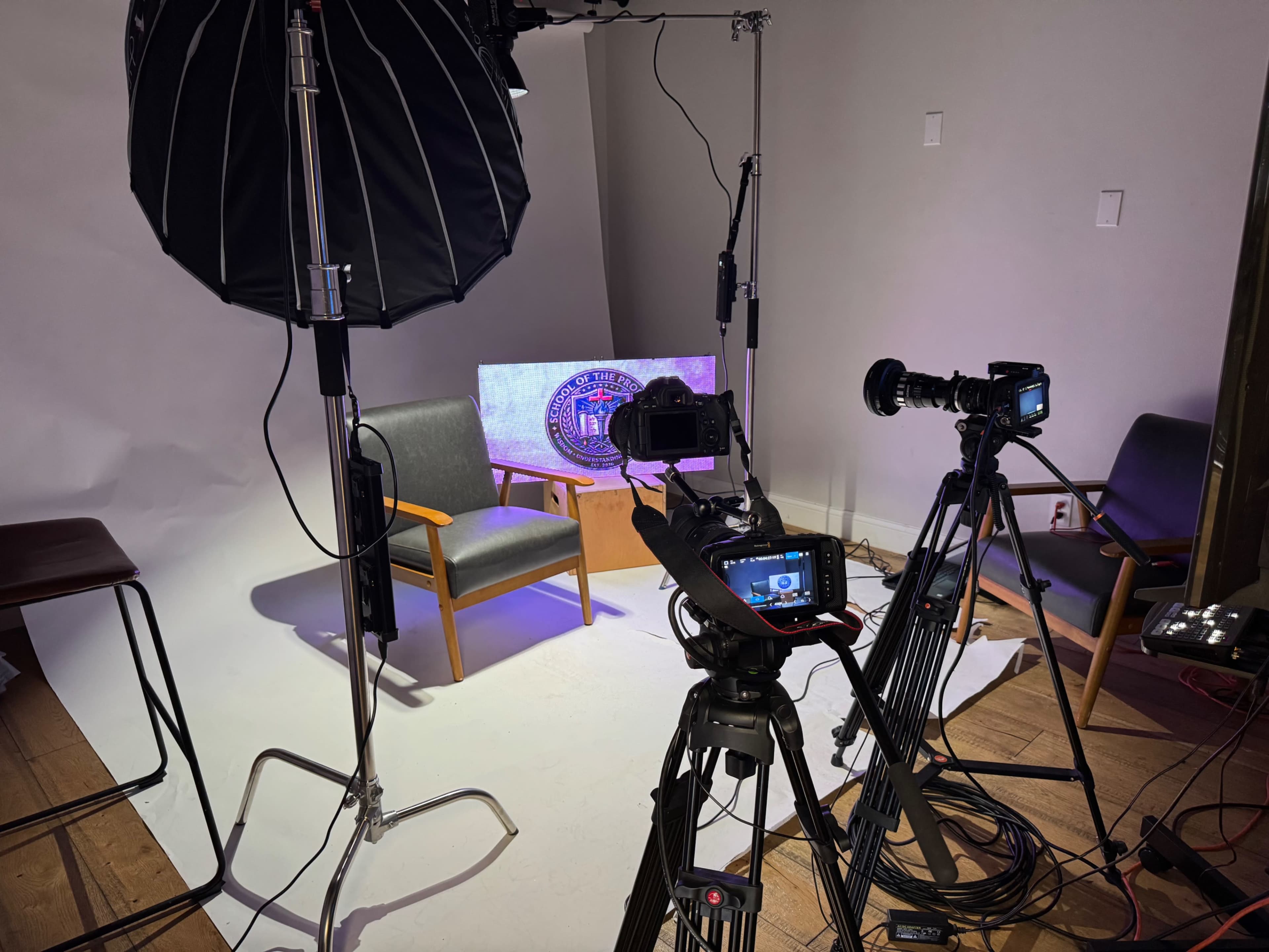 The image shows a setup for a video shoot, featuring two chairs, multiple cameras, and lighting equipment on a backdrop.