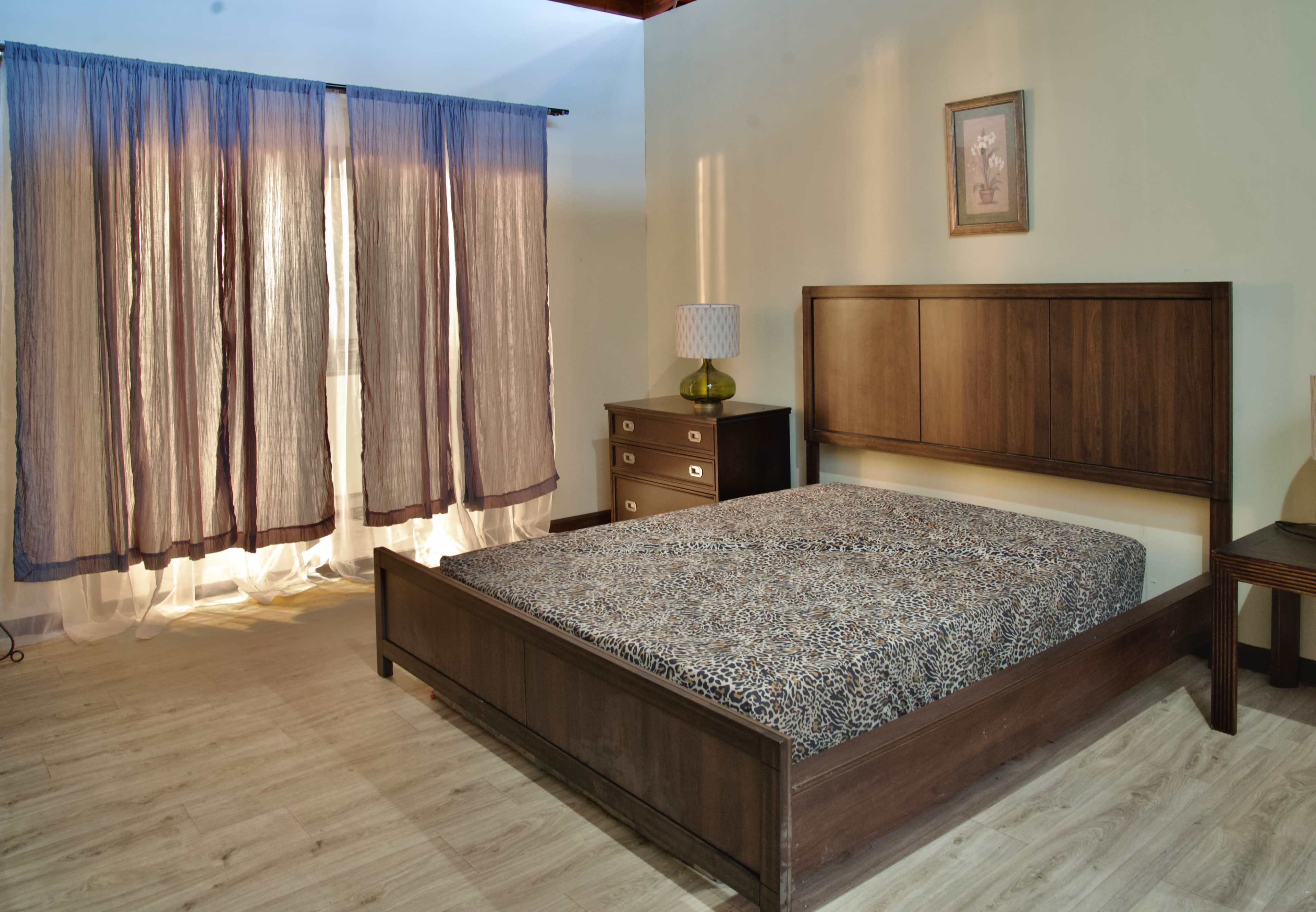 A bedroom with a wooden bed frame, a patterned mattress, a nightstand, and curtains letting in natural light.